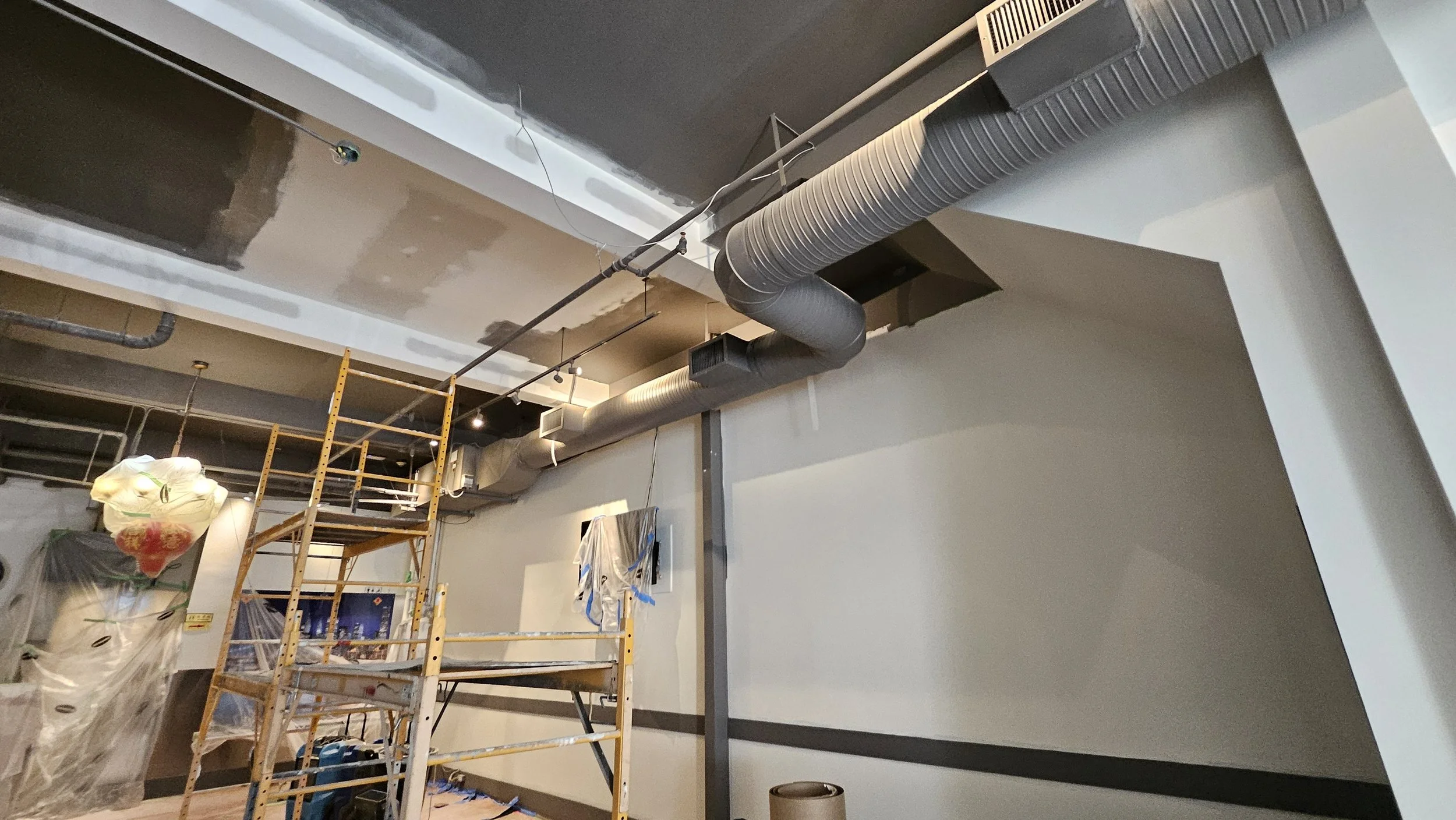 Interior of a building under construction, with partially painted walls, exposed ductwork, scaffolding, and construction materials.