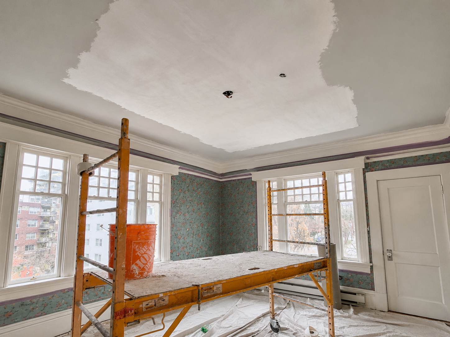 Room under renovation with a suspended scaffolding, three large windows, floral wallpaper, and a partially painted ceiling.