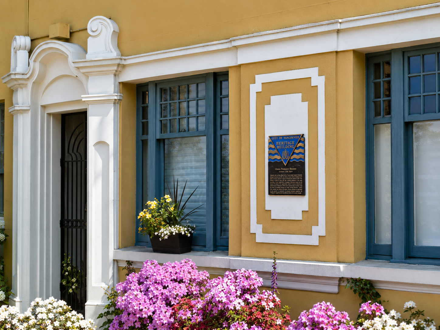 Exterior of a historic building with yellow walls, blue window frames, and a sign that reads 'City of Vancouver Heritage Building'. Flowering plants in front.