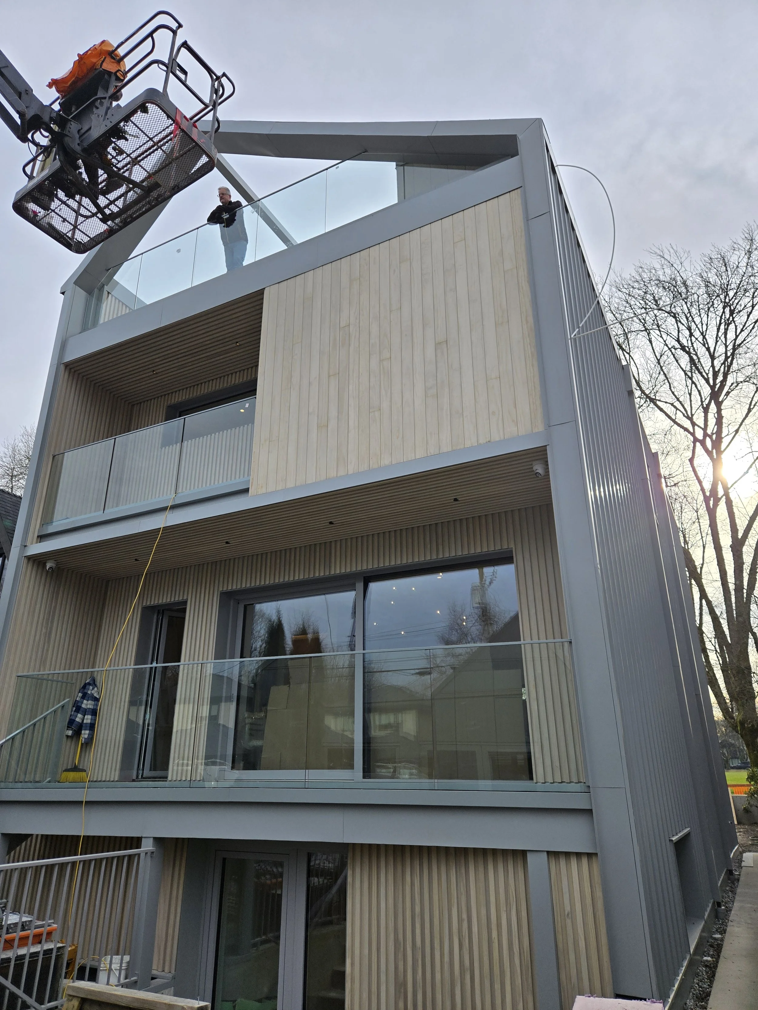 A multi-story modern building with glass balconies and vertical wooden paneling, seen from the ground looking upward. A construction lift with a person inside extends towards the top of the building, and the sky is overcast with bare trees visible on the right side.