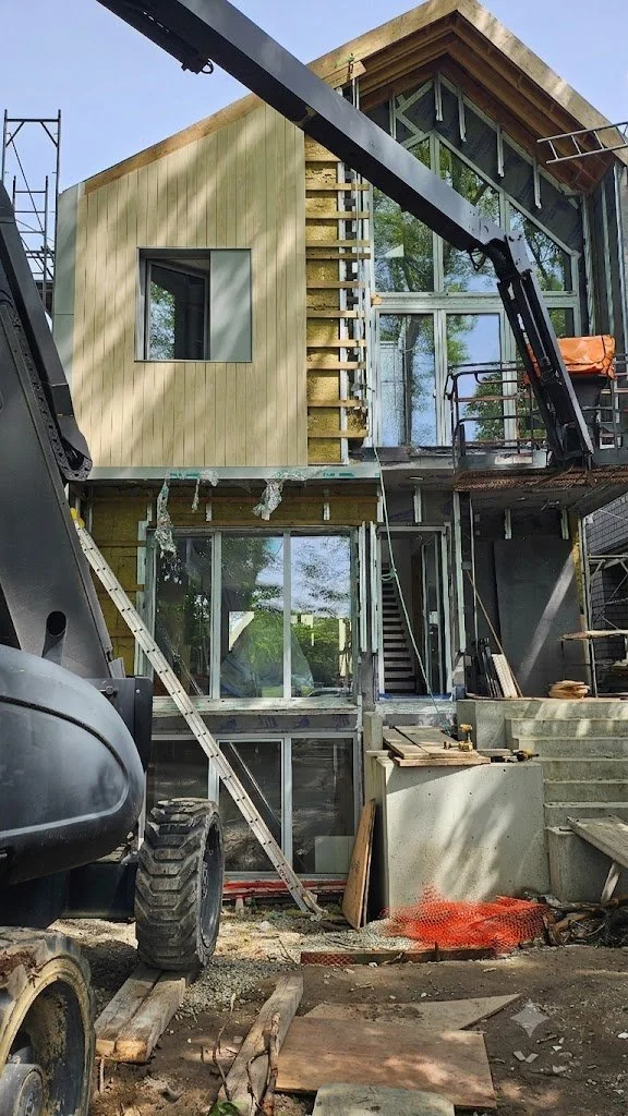 Under construction modern multi-story house with large glass windows and wooden exterior panels, construction equipment and materials surrounding it.