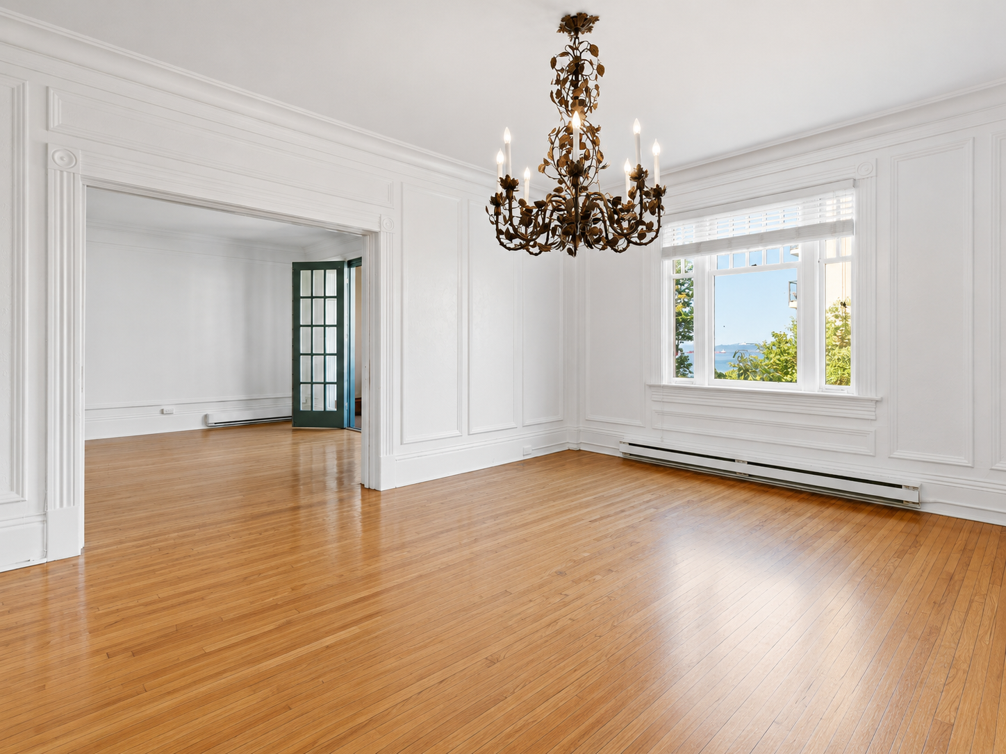 Bright, empty room with white walls, hardwood floors, a chandelier, large window with outside view, and a doorway leading to another room.