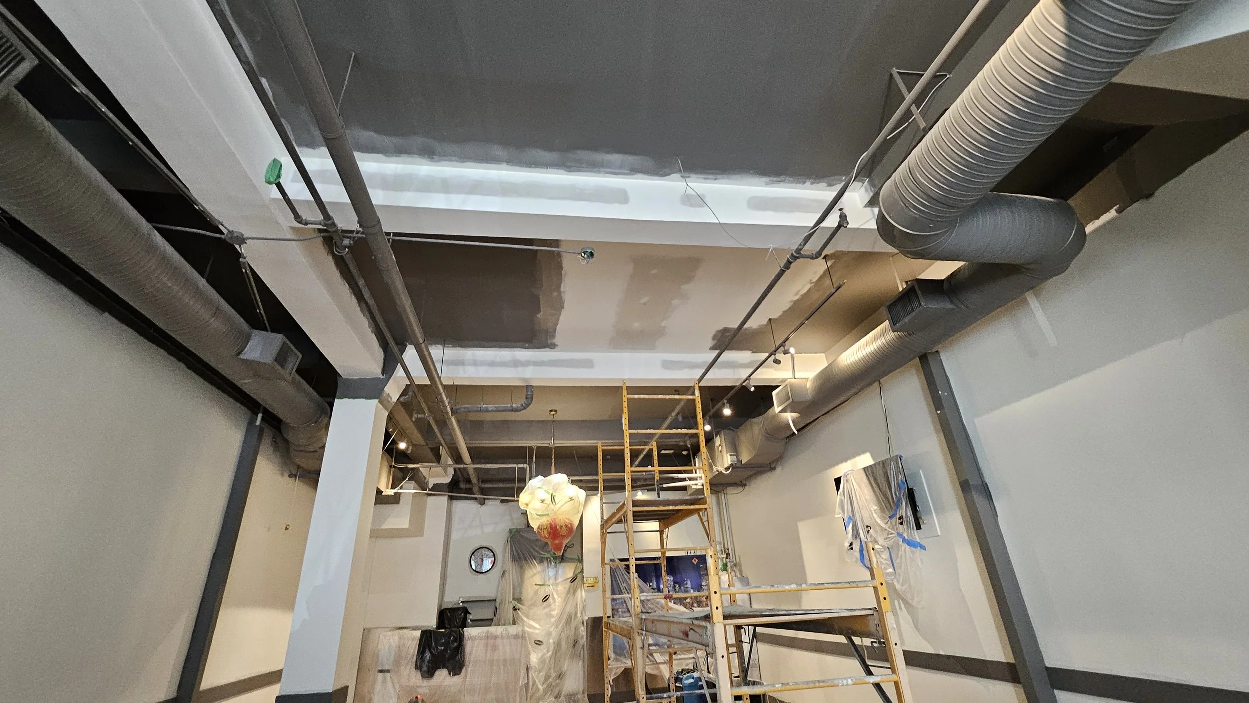 Construction or renovation scene with exposed ceilings, ducts, pipes, a ladder, and construction materials in an interior space.