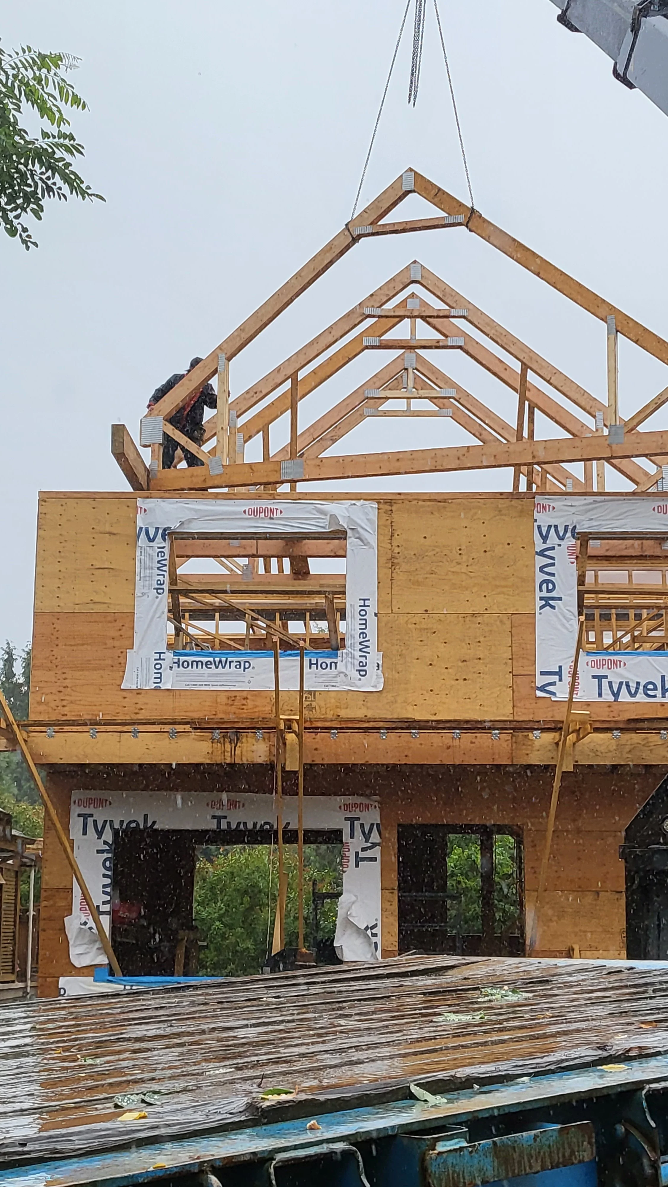 Construction site of a residential building with wooden framing, window openings, and scaffolding, in rainy weather.