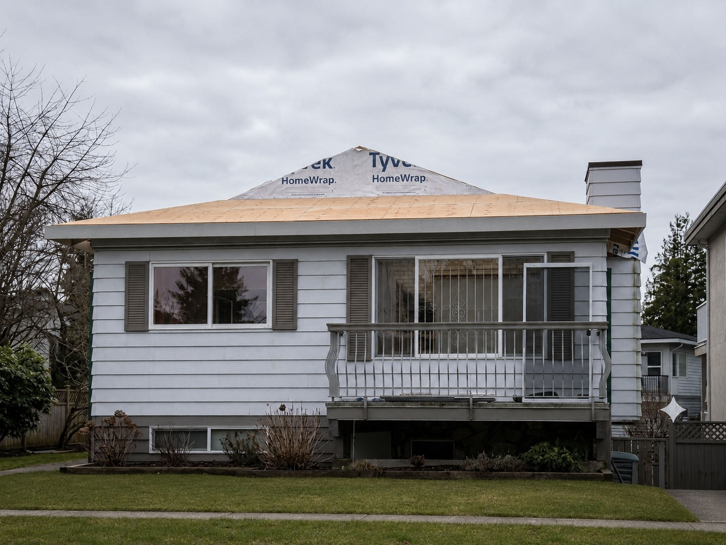 Front of a house under renovation with new roof shingles, white siding, and a small wooden deck with a metal railing.