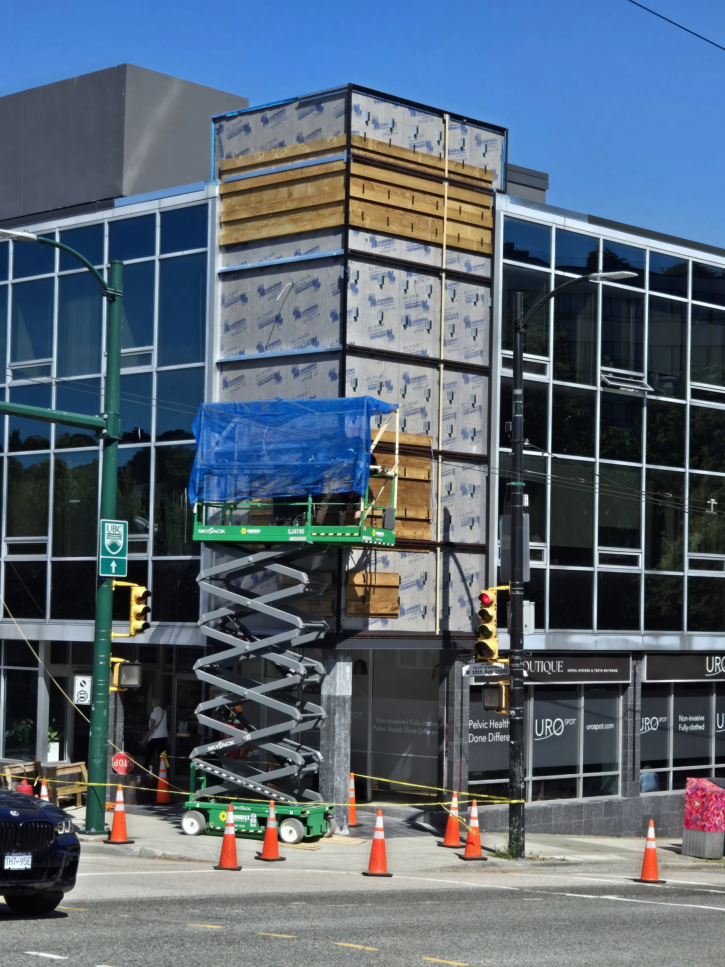 Building under construction with glass windows, construction lift with wooden pallets, traffic cones, and a stop sign on the street.