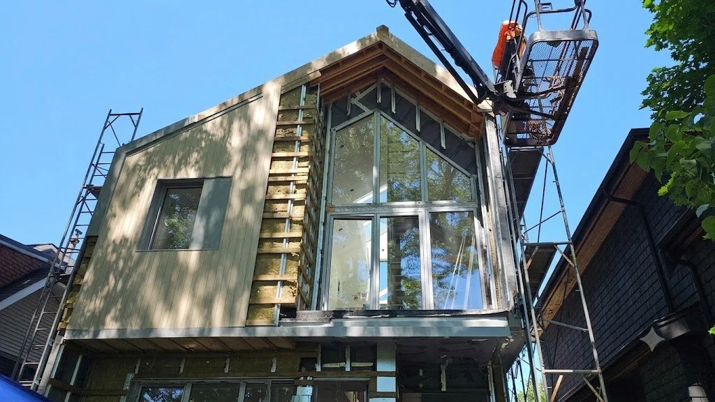 A house under construction with a metal frame and glass windows, on scaffolding with a cherry picker lift nearby on a sunny day.