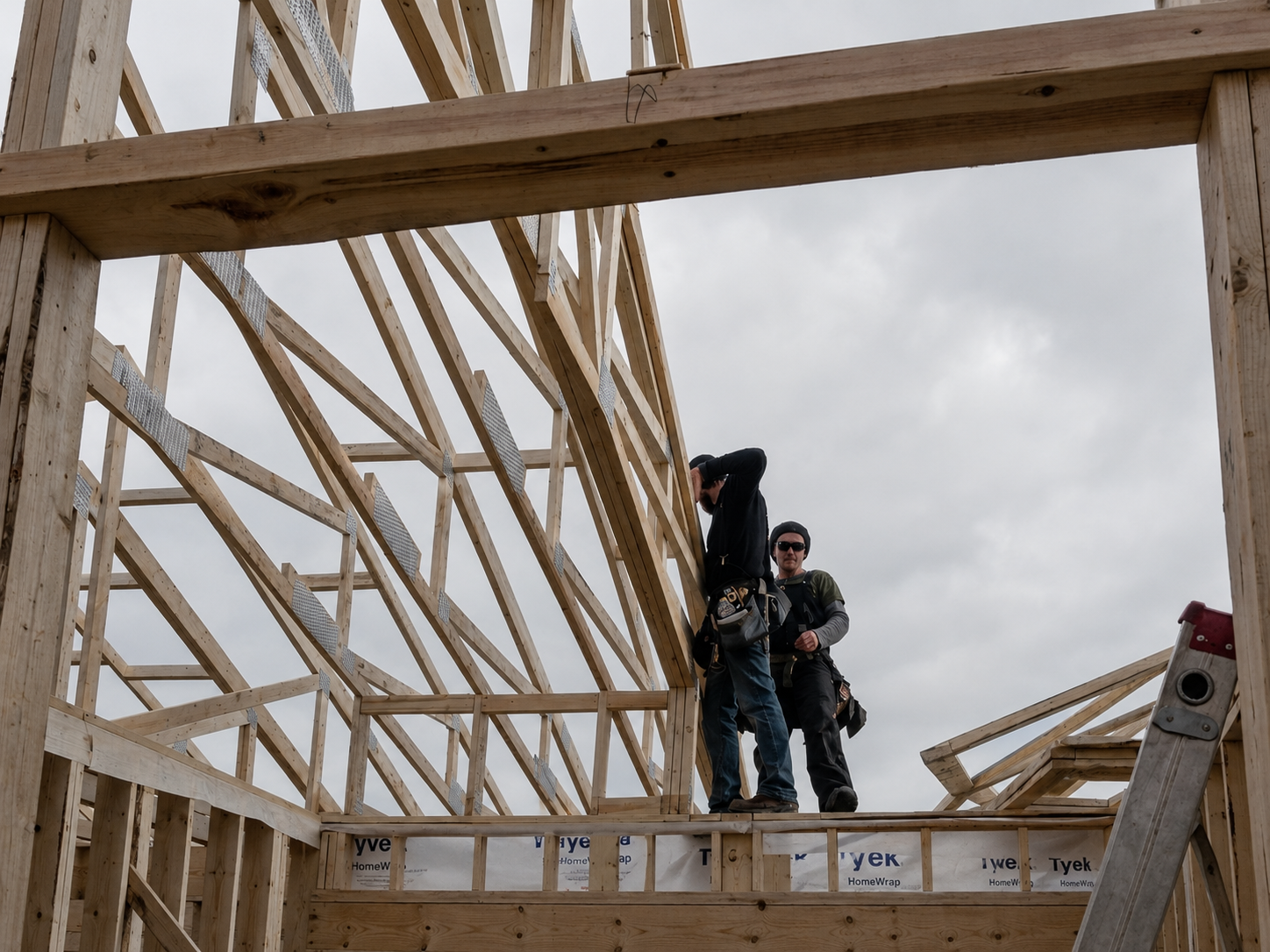 Two construction workers standing inside a wooden framing structure on a cloudy day.