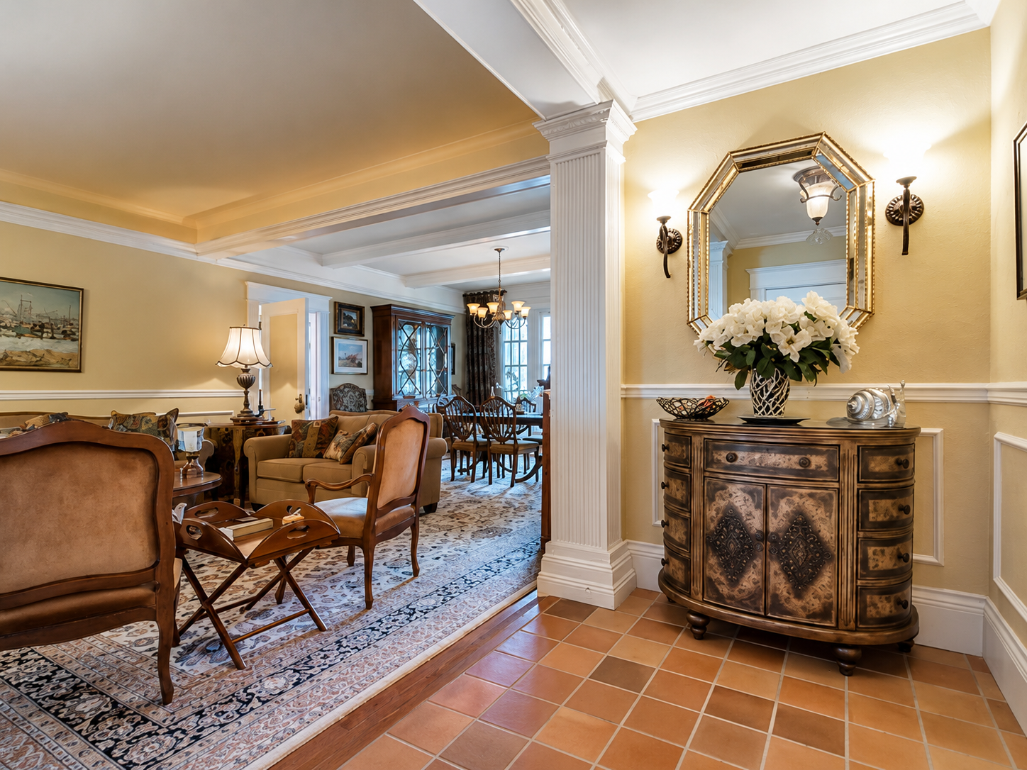 View of a traditional living room with beige walls, classic furniture, a chandelier, and a dining area in the background. An ornate wooden sideboard with flowers and a mirror is in the foreground.