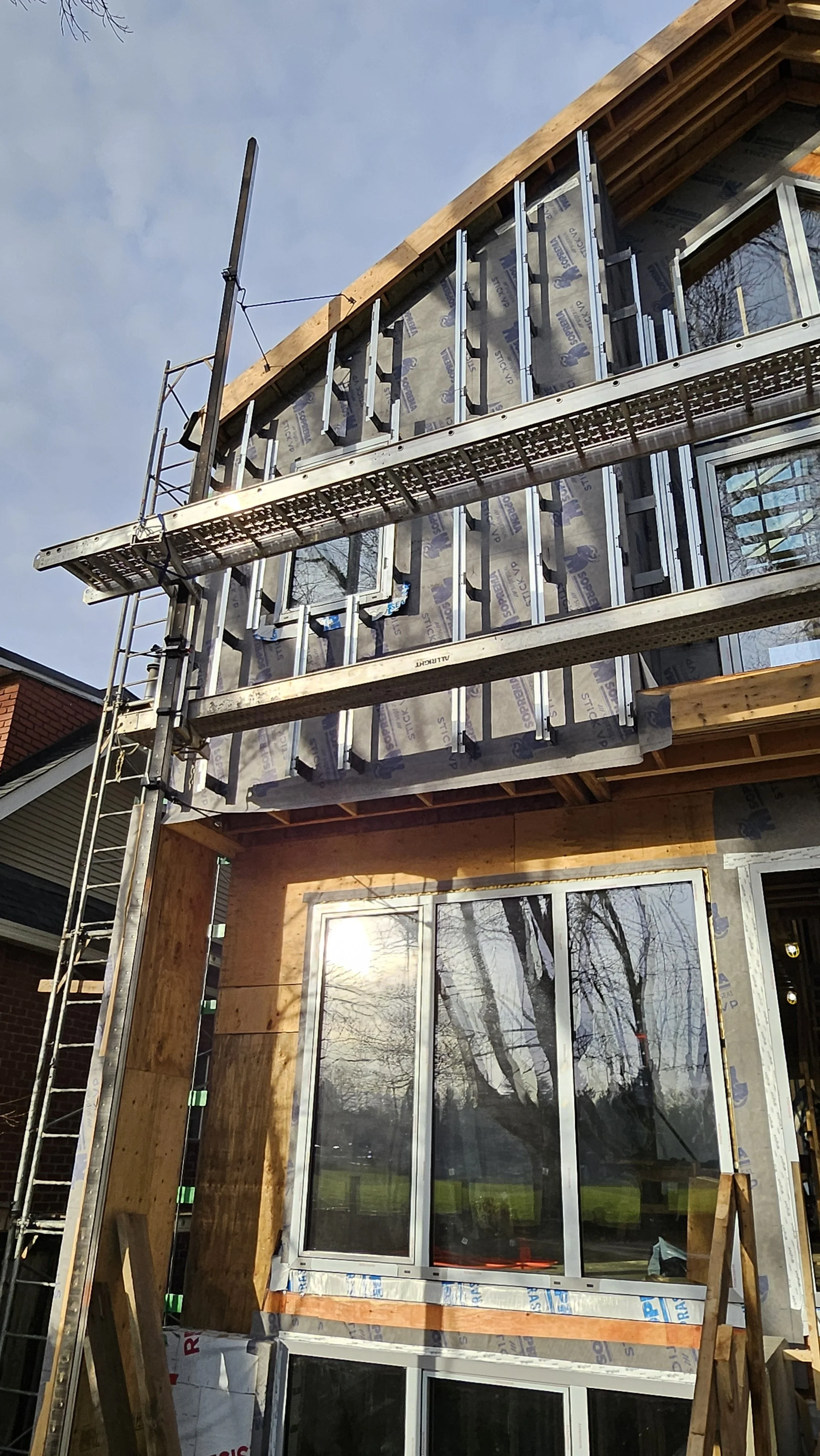A house under construction showing exposed framing, siding, and windows, with scaffolding on the left side and a blue sky in the background.