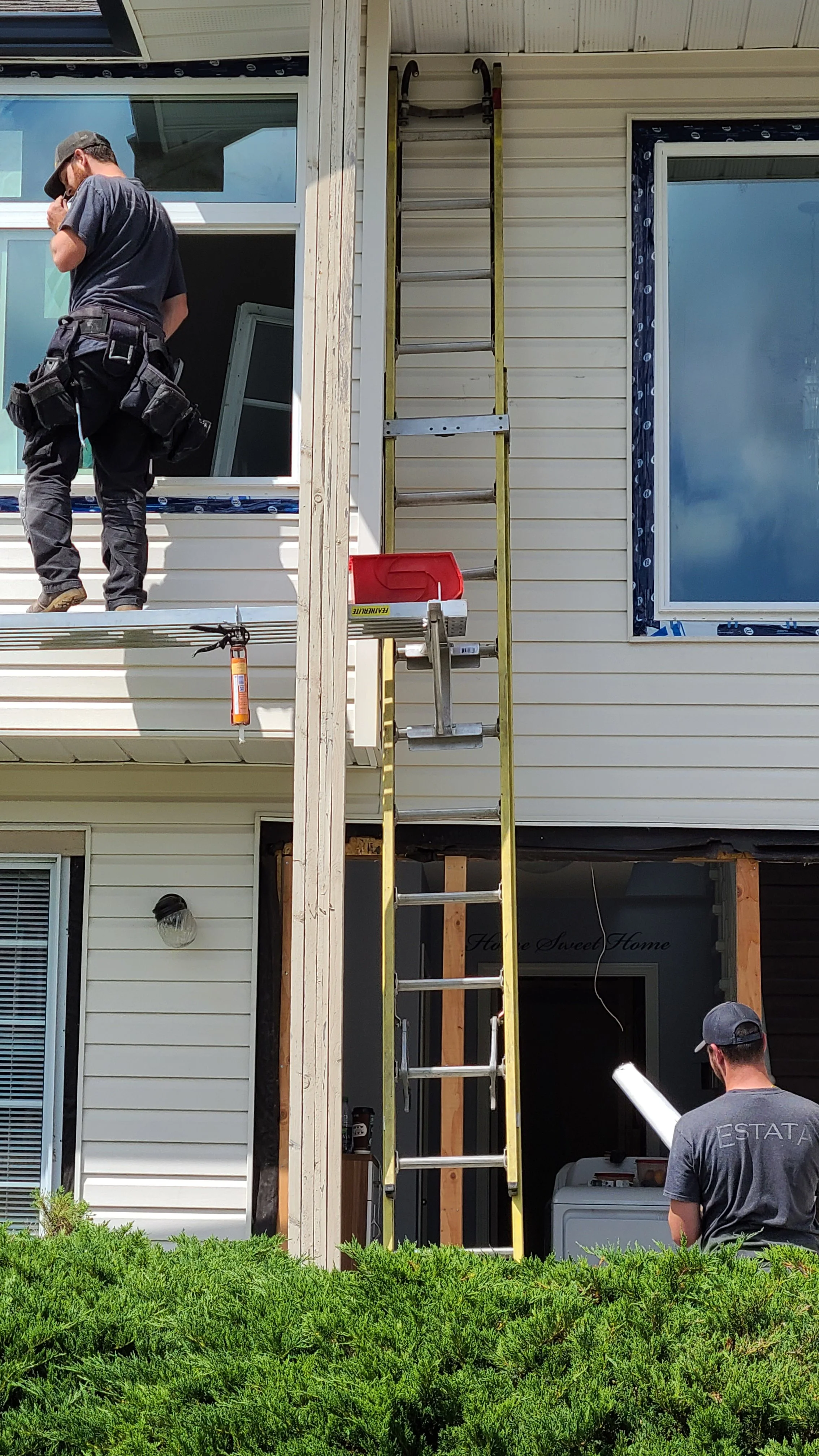 Workers are installing siding on a house. One person is on the balcony near the window, and another person is standing on the ground holding a rolled-up blueprint. A ladder is positioned against the house, and various tools are nearby, with all the house's exterior panels partially installed.
