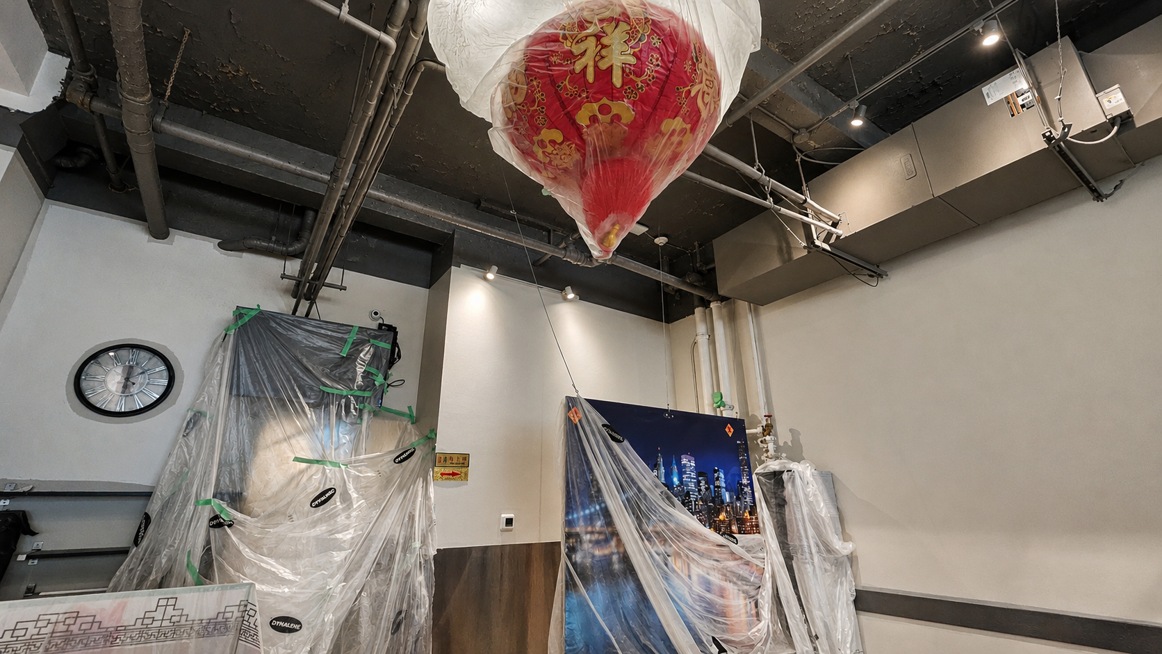 Interior room with a large red Chinese lantern hanging from the ceiling, plastic protective coverings on objects, a wall clock, and a cityscape picture, with exposed industrial pipes and equipment on the ceiling.