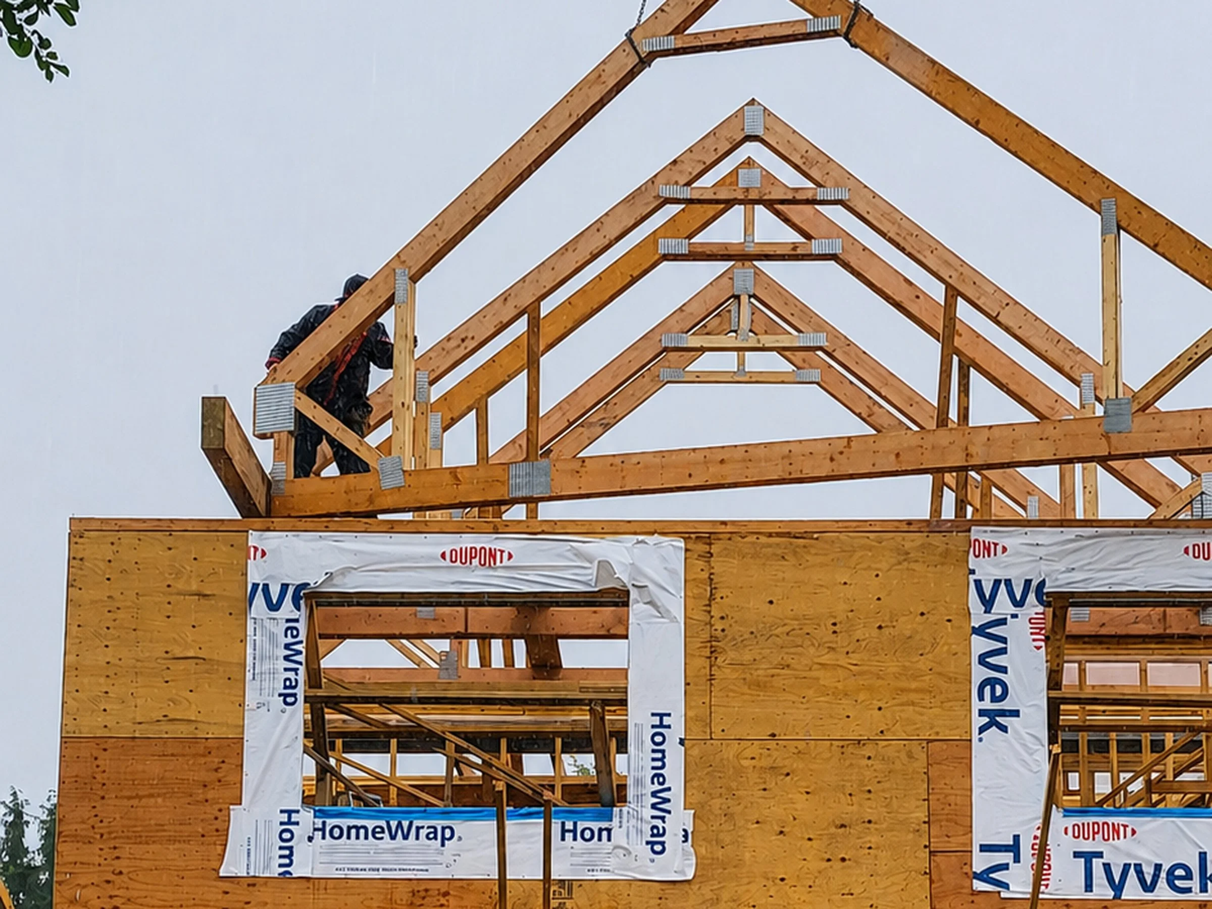 Wooden framing of a house under construction, with visible roof trusses and walls wrapped in HomeWrap, and partially visible signs for DuPont and HouseWrap.