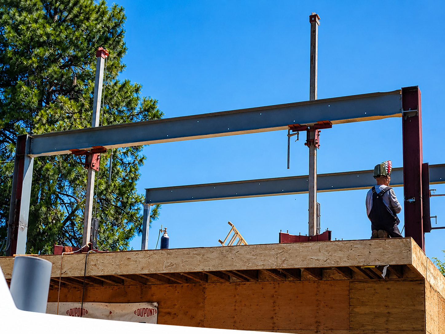 Construction worker standing on a partially built wooden structure, with steel beams and construction materials around, under a clear blue sky and a leafy green tree.