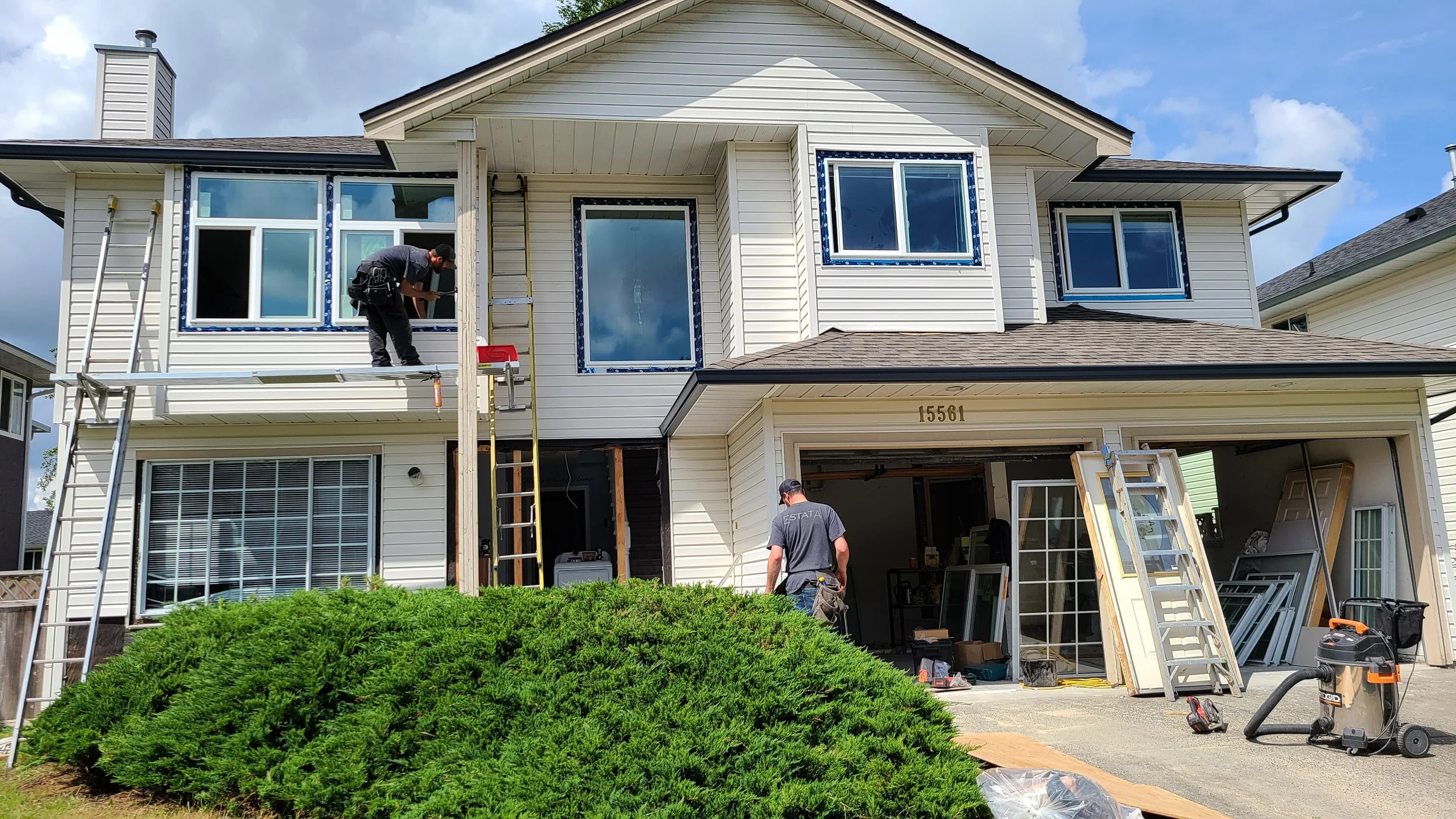 Construction workers are installing new windows on a two-story house. Some workers are on a scaffold, and others are on the ground, with tools and materials around them. The house has light-colored siding and a garage with an open door. The sky is partly cloudy.
