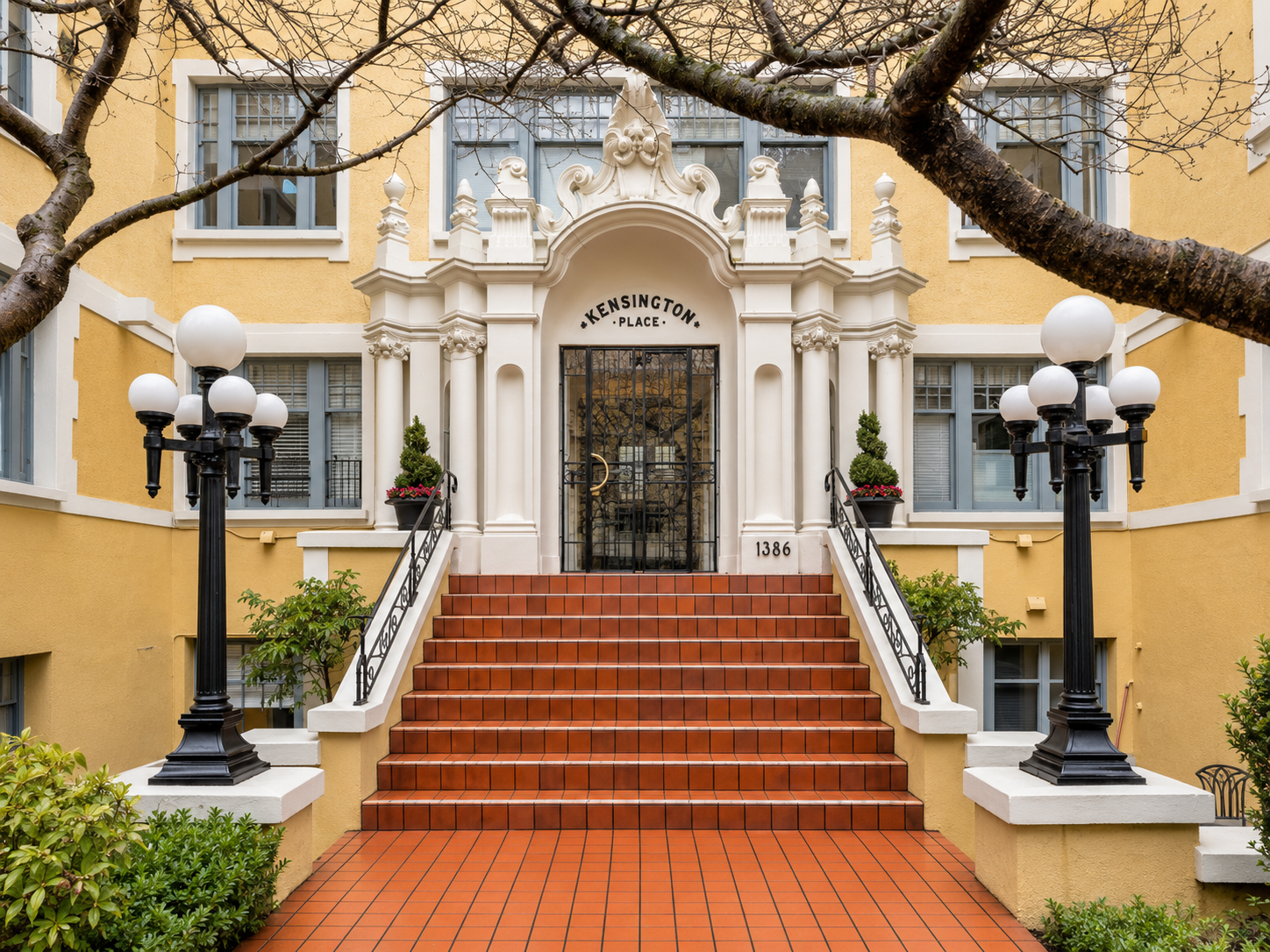 Entrance to building named Kensington Place featuring red brick stairs, black wrought iron railings, white columns, decorative archway, black gate, two black lampposts, potted plants, and yellow exterior walls.