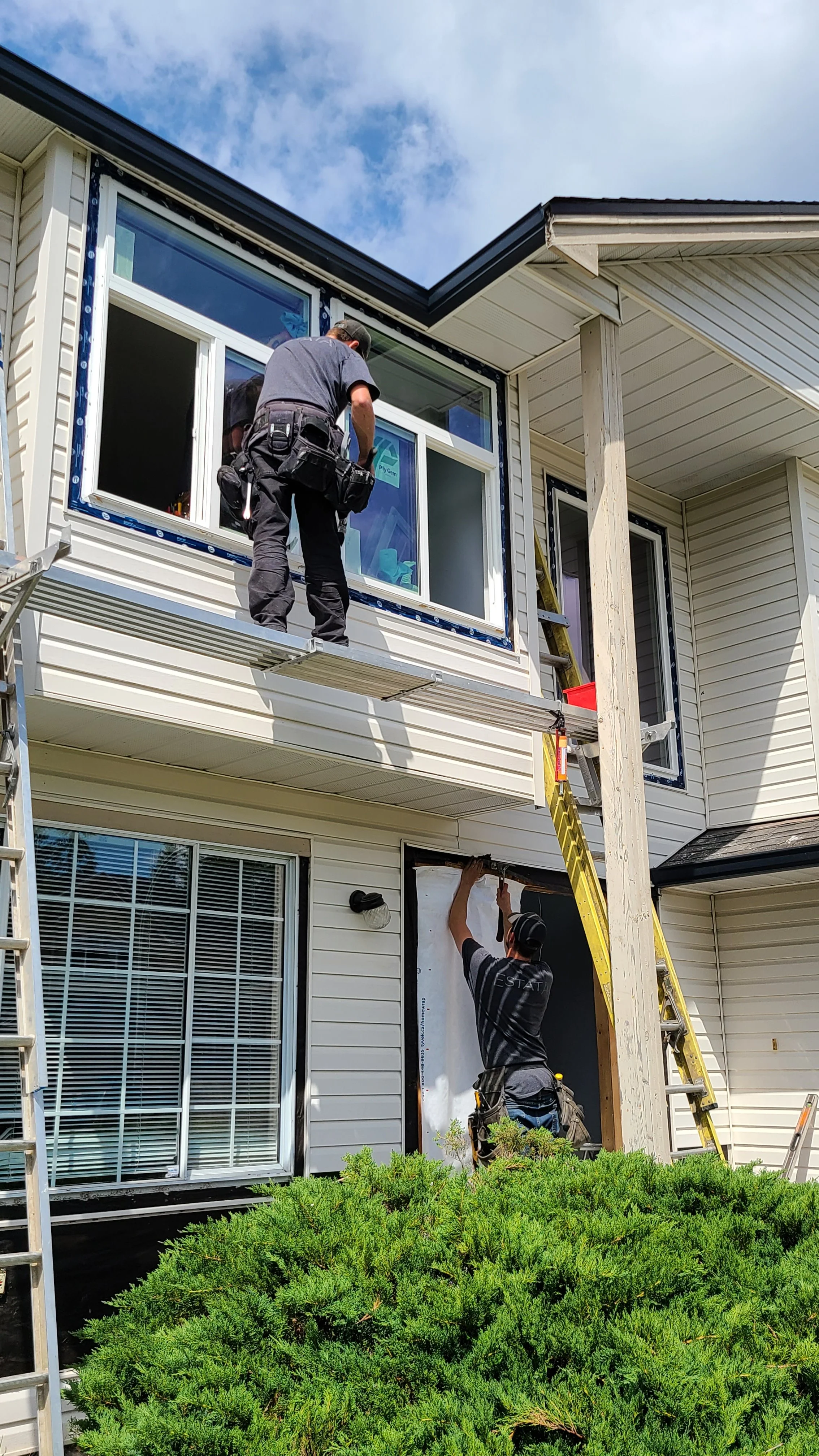 Workers installing a window on the second story of a house, with ladders and tools, in a residential neighborhood under a partly cloudy sky.