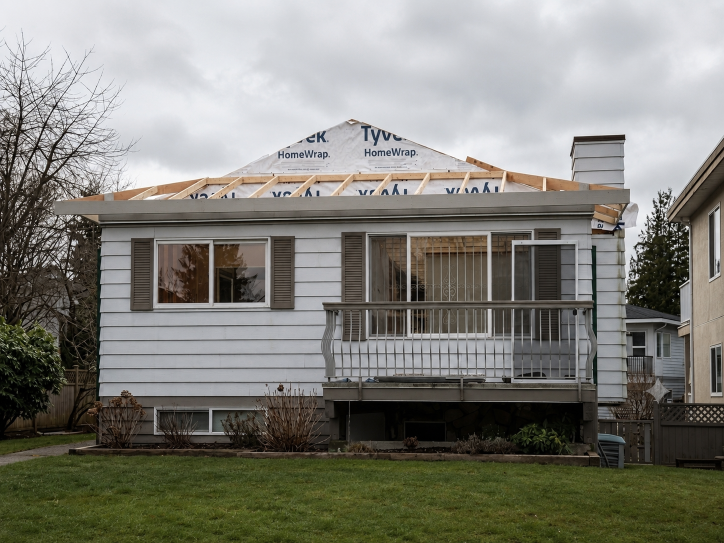 A house under renovation with exposed roof trusses, white siding, a small balcony with a white railing, and a backyard with green grass and shrubs. The sky is overcast.