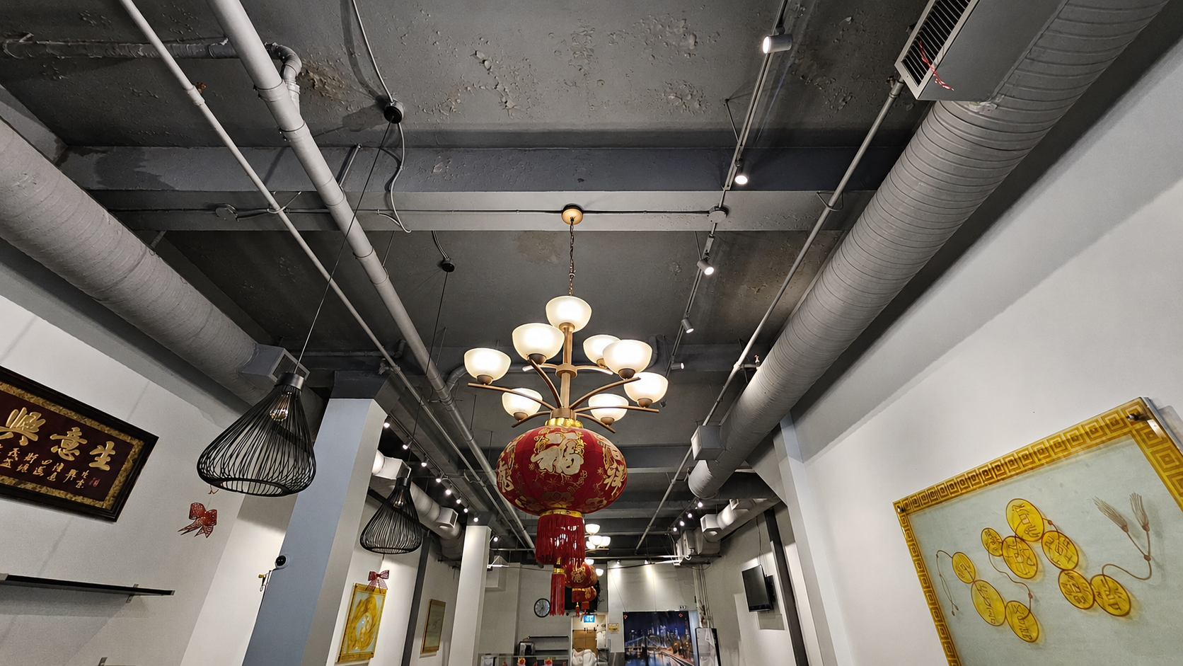 Interior of a room with a decorative ceiling featuring exposed pipes, a chandelier with multiple globes, and hanging lanterns. Wall art and a clock are visible, with a mix of modern and traditional decor.