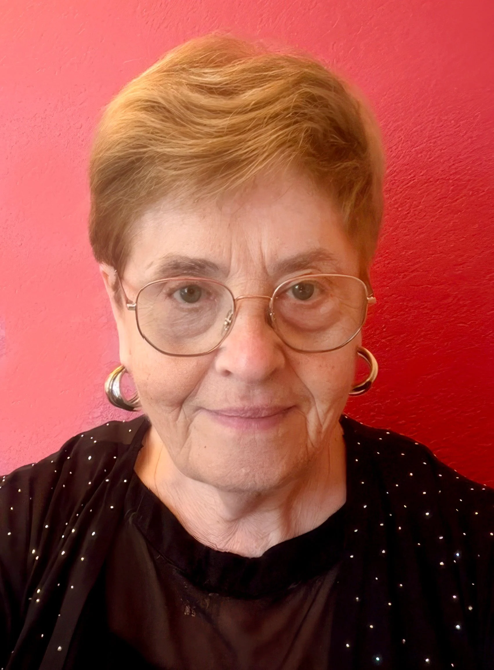 Close-up of a woman with short, reddish hair, wearing glasses and earrings, smiling, against a pink textured wall and wearing a black top with small white polka dots.