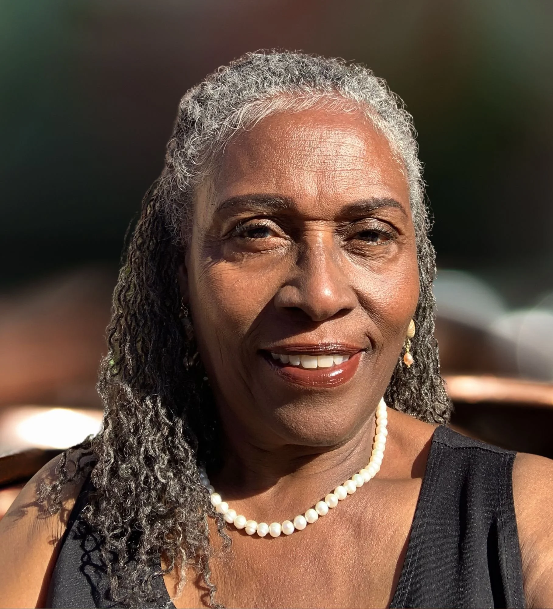 Close-up of a smiling woman with gray curly hair, wearing pearl necklace and earrings, in a black sleeveless dress.