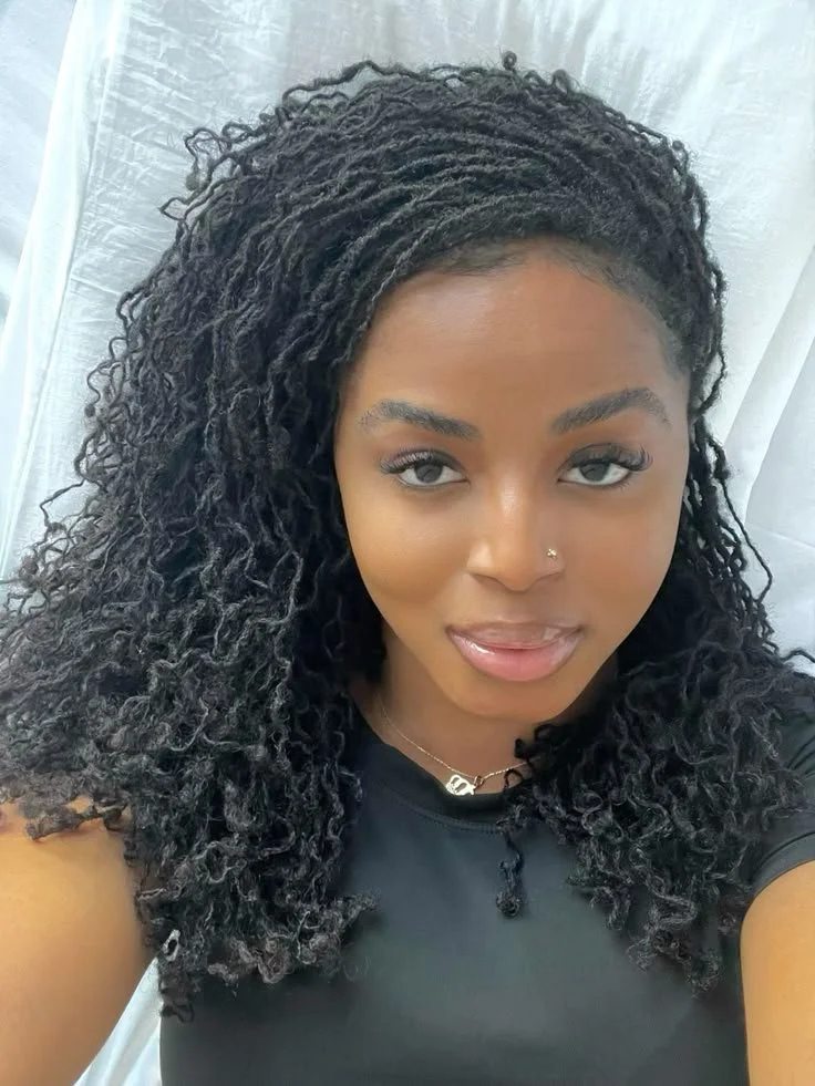 A woman with curly black hair, light makeup, a nose piercing, and wearing a black top and jewelry, looking at the camera with a slight smile, standing against a white background.