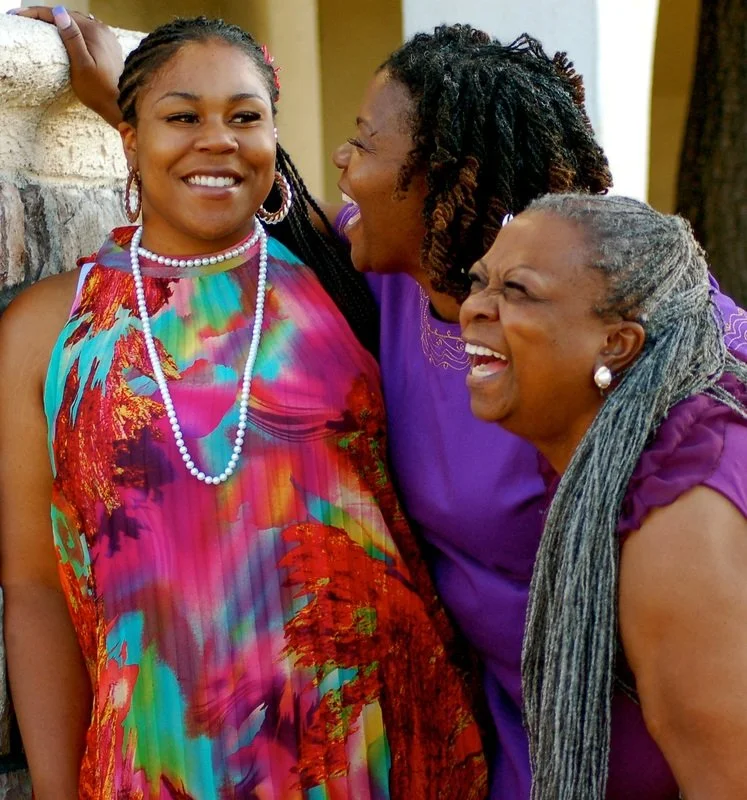 Three women laughing and smiling, standing close together outdoors. The woman on the left wears a colorful dress with a floral pattern and pearl necklace, the woman in the middle wears a purple top, and the woman on the right wears a purple shirt with gray braided hair.