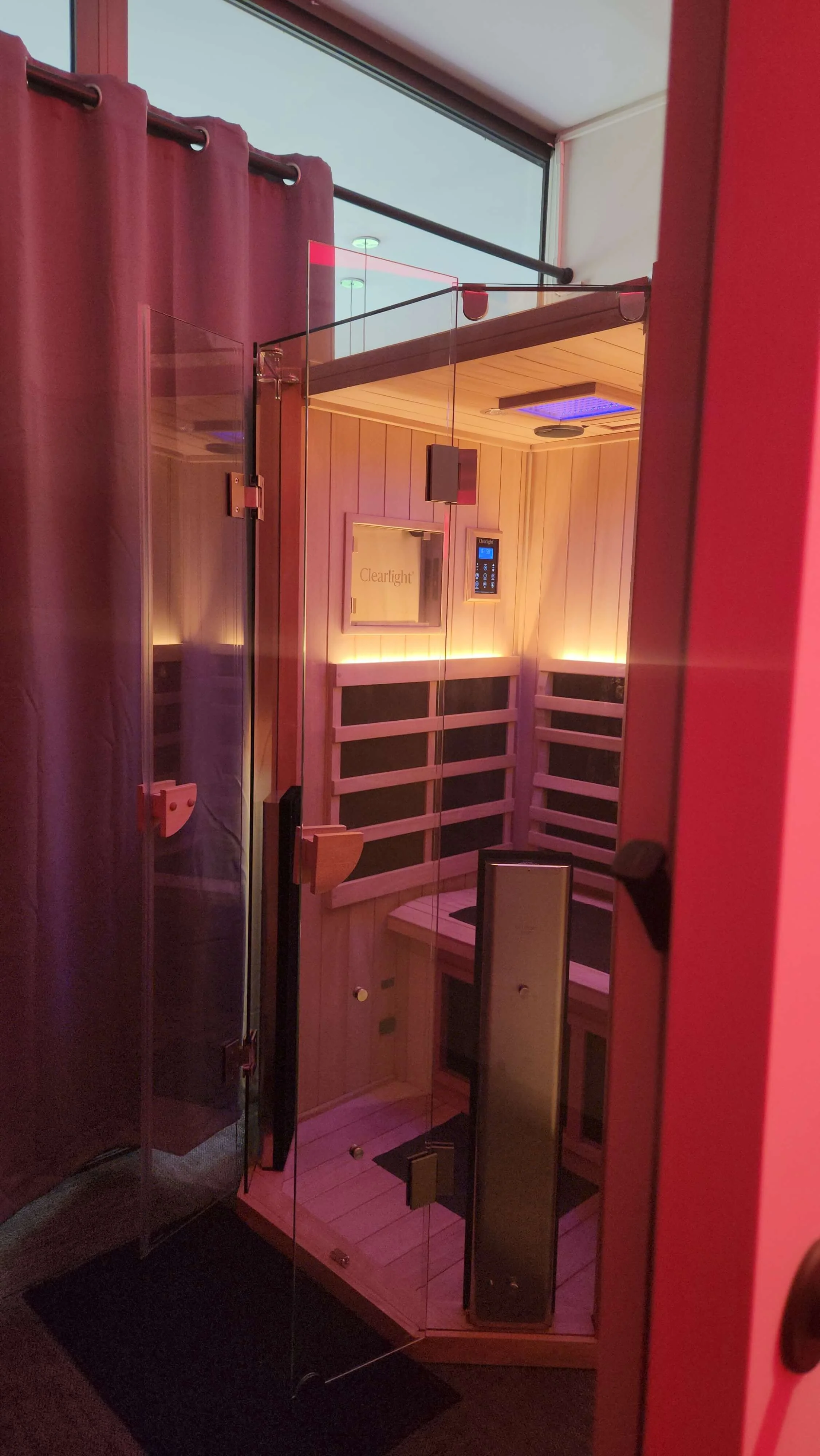 Interior of a sauna with wooden walls and benches, glass door, and a control panel on the wall. The lighting is warm with a purple hue on the ceiling.