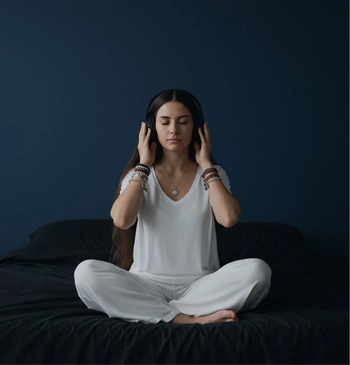 Woman sitting cross-legged on bed, wearing white pajamas and headphones, with eyes closed in a meditative pose against a dark blue wall.