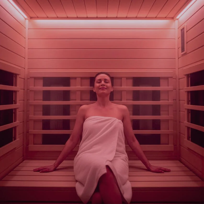 A woman wrapped in a towel sitting inside a wooden infrared sauna emitting a red glow.