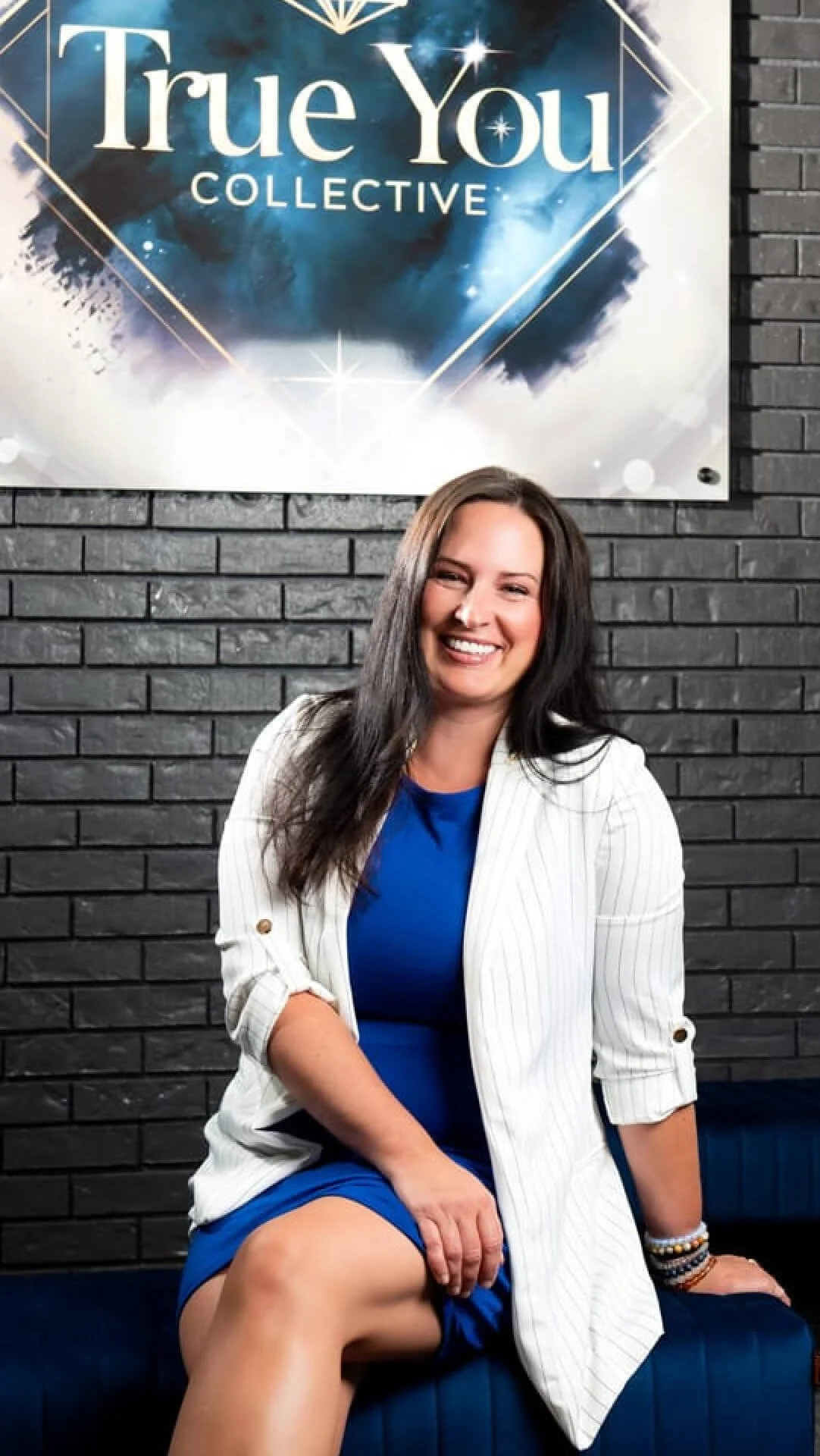 A smiling woman with long dark hair, wearing a white striped blazer and a blue dress, seated on a dark blue bench in front of a black brick wall. Behind her, there is a large sign that reads 'True You Collective' with a cosmic background and geometric star design.