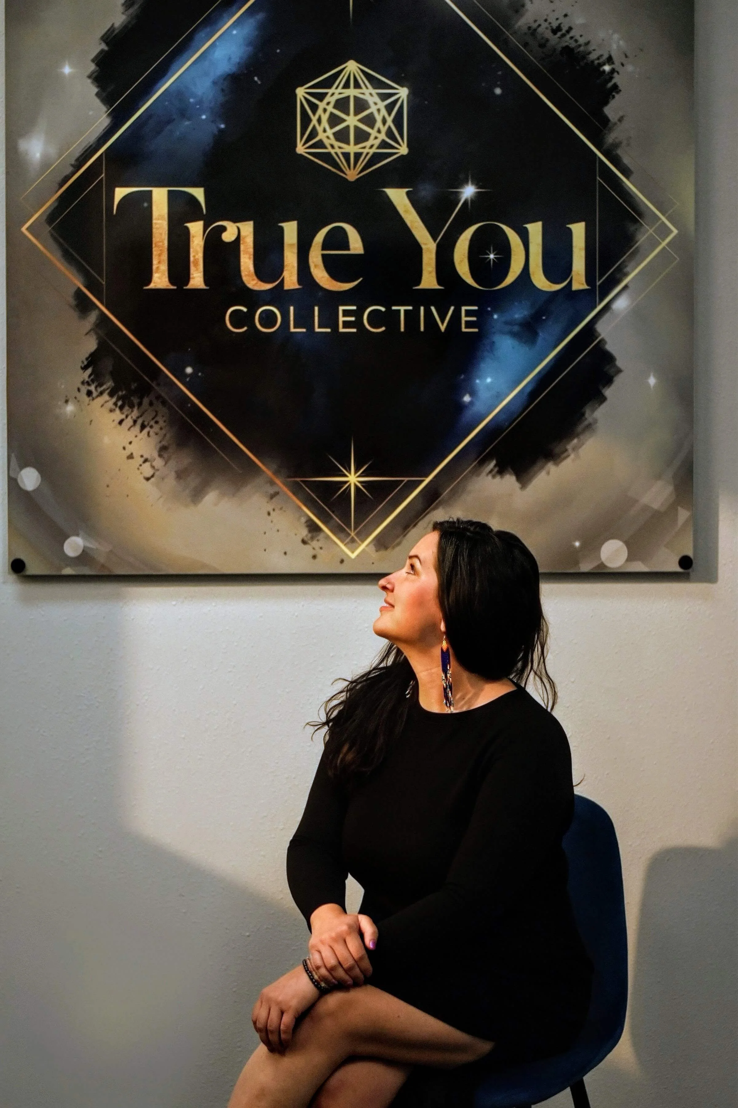 A woman with long dark hair, wearing earrings, a black long-sleeve top, and a bracelet, is sitting on a blue chair, looking up and to her left in front of a wall with a large poster that reads 'True You Collective' with a geometric design above.