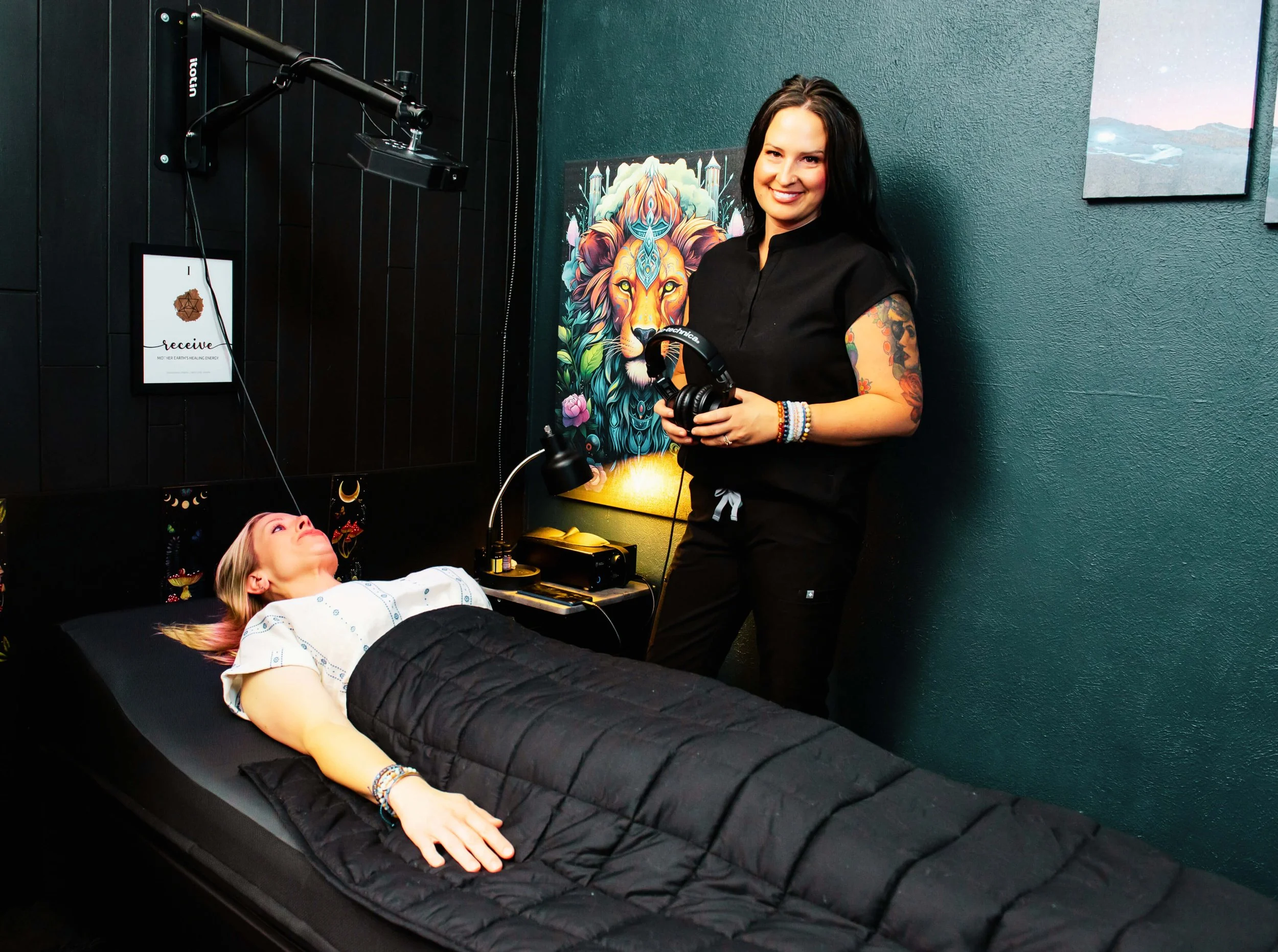 A woman with long dark hair and tattoos on her arms is smiling while holding a pair of headphones, standing beside a person lying on a treatment table with a black quilted blanket. The person on the table has blonde hair, is wearing a patterned shirt, and appears relaxed with eyes closed.