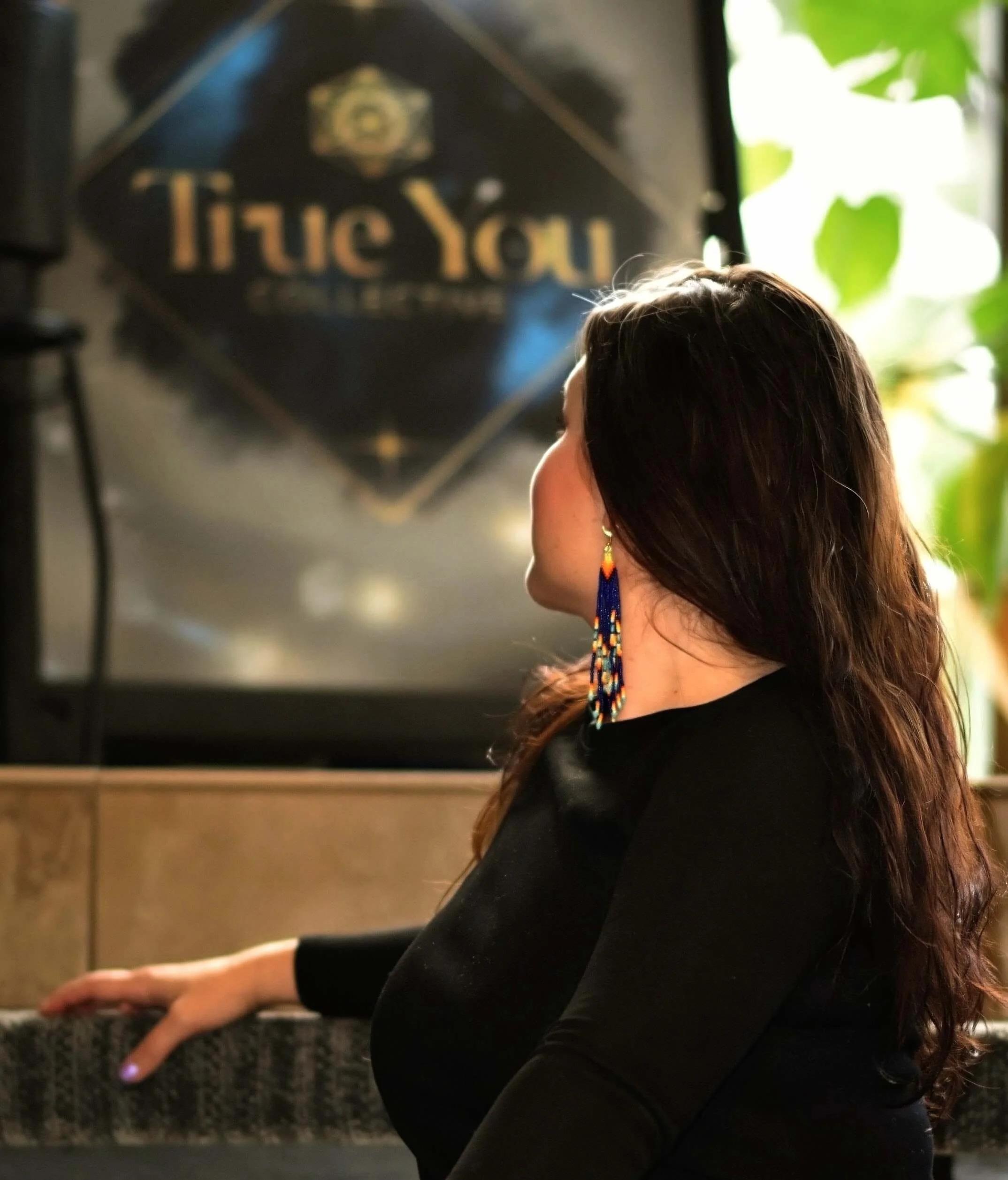 A woman with long brown hair and colorful beaded earrings is playing a piano, with a window and greenery in the background. Blurred wall art with the words 'I love you' is visible in the background.