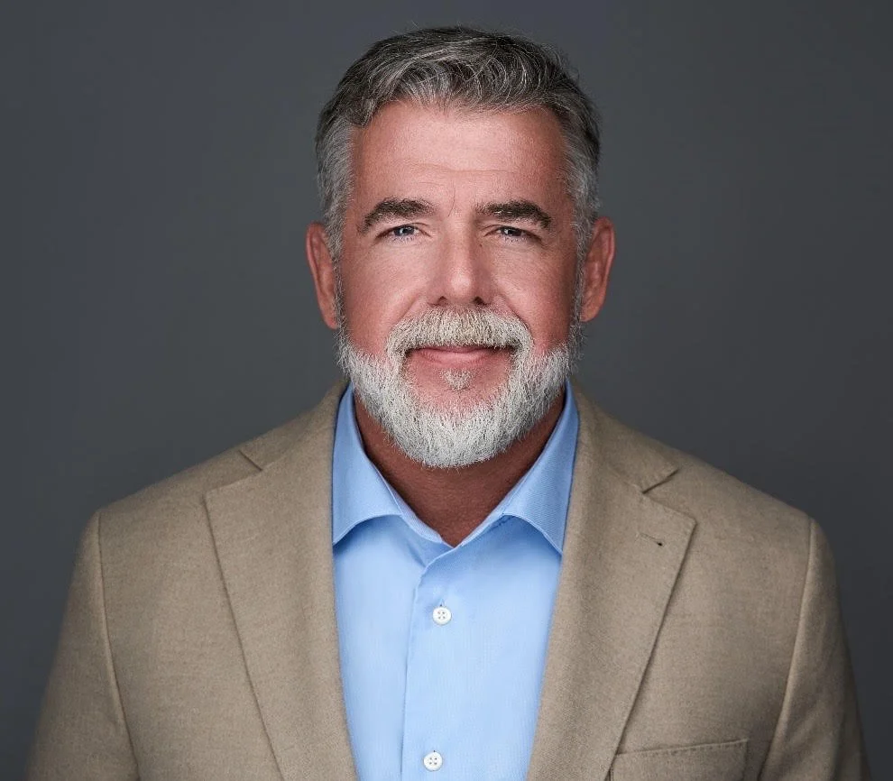 Headshot of a senior man with gray hair and a beard, wearing a tan blazer and a blue dress shirt, smiling against a gray background.