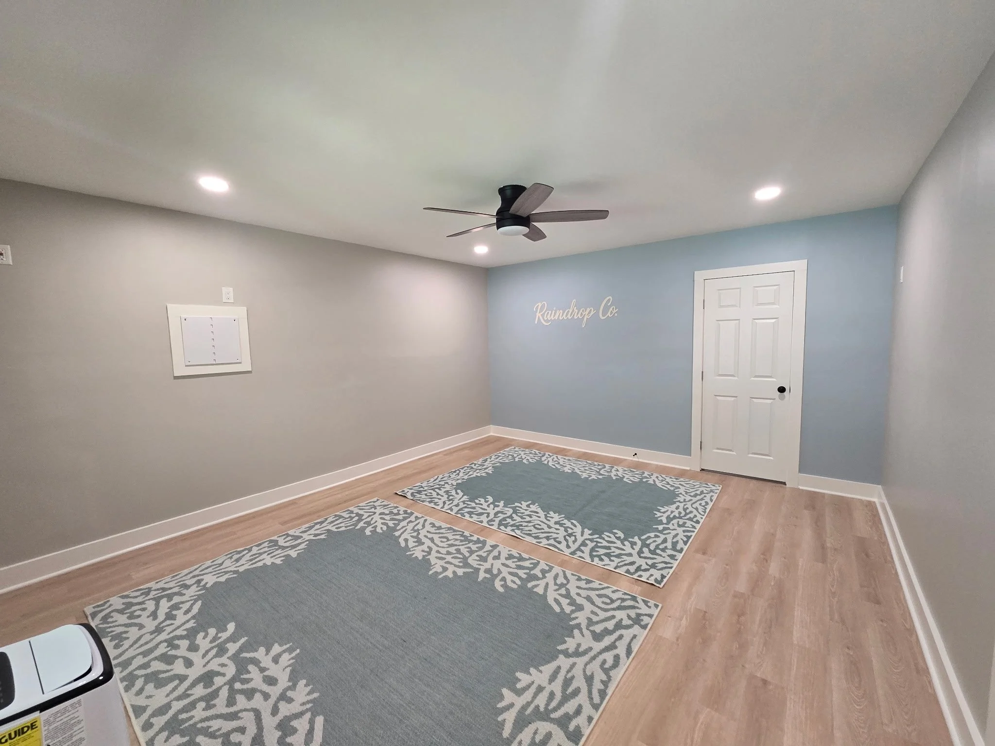 A room with beige walls, a white door, hardwood floors, a ceiling fan, and three patterned area rugs.