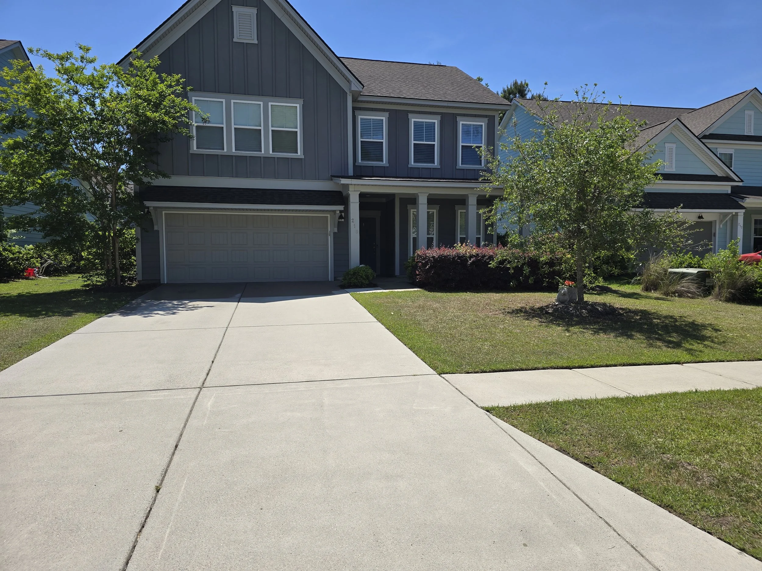 A two-story suburban house with gray and blue siding, a garage, and a front porch. There are trees and shrubs in the front yard, and a clear blue sky overhead.