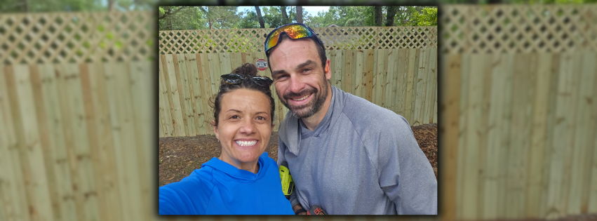 A smiling woman and man taking a selfie outdoors with a wooden fence in the background.