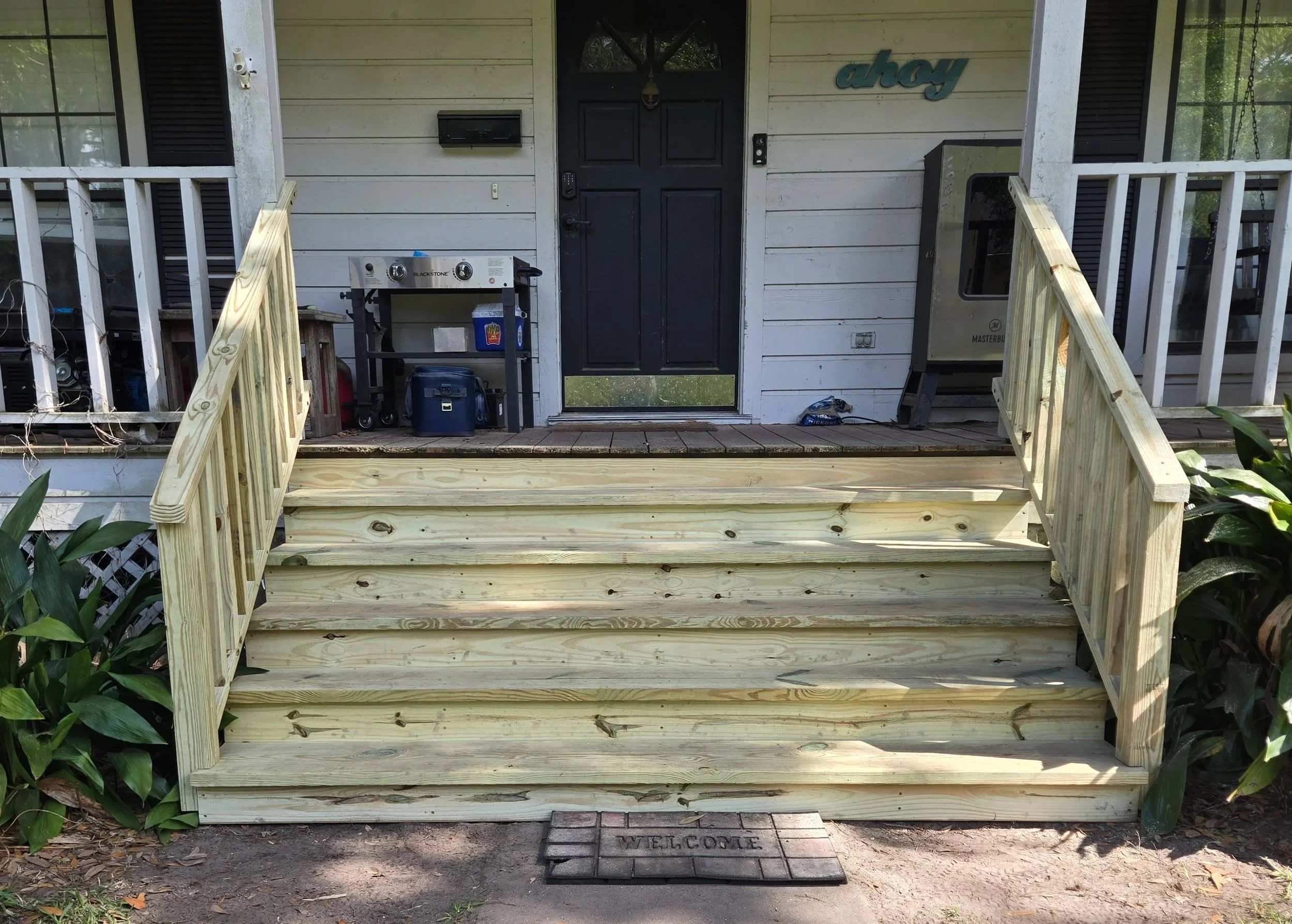 Front porch of a house with a set of new wooden stairs, a black door, a welcome mat, outdoor grill, mailbox, and a house sign reading 'ahoy'.