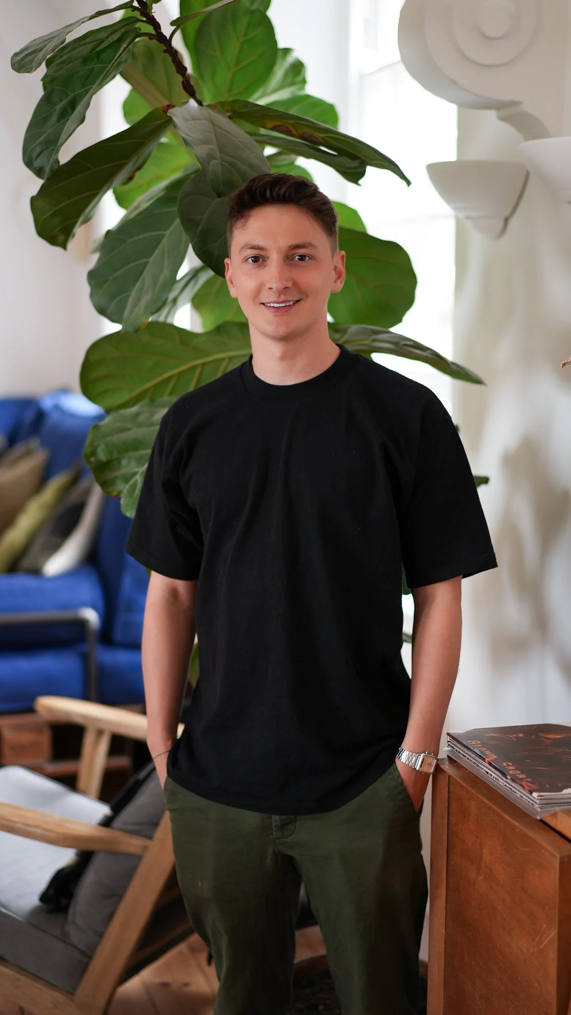 Young man standing indoors near a large green plant, smiling, wearing a black T-shirt, green pants, and a watch, with a blue chair and some books on a wooden table in the background.