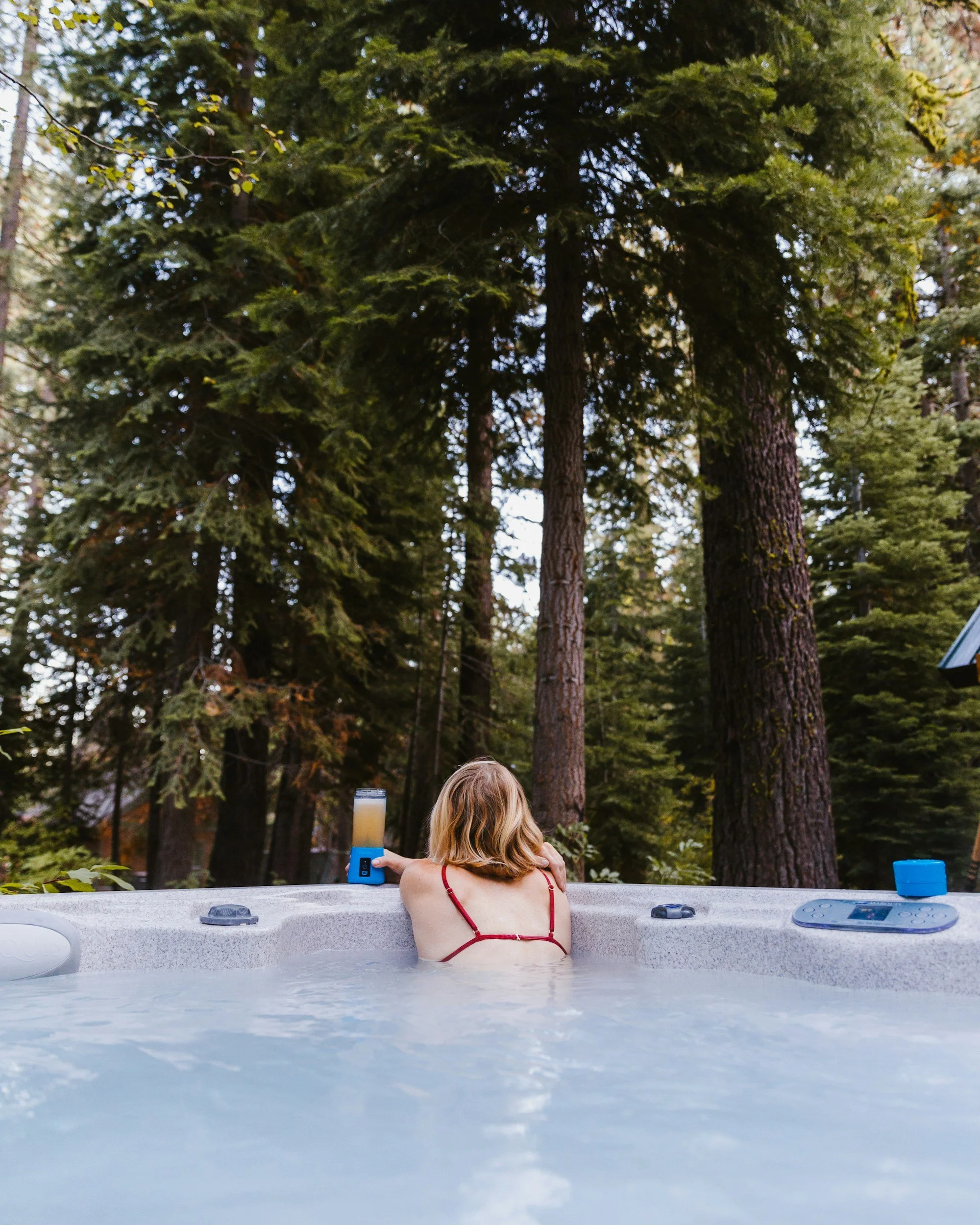 A woman with blonde hair in a red swimsuit is sitting in a hot tub outdoors, facing away. She has a drink with a candle on the side table nearby. Trees surround the hot tub, and the setting appears to be a forested area.