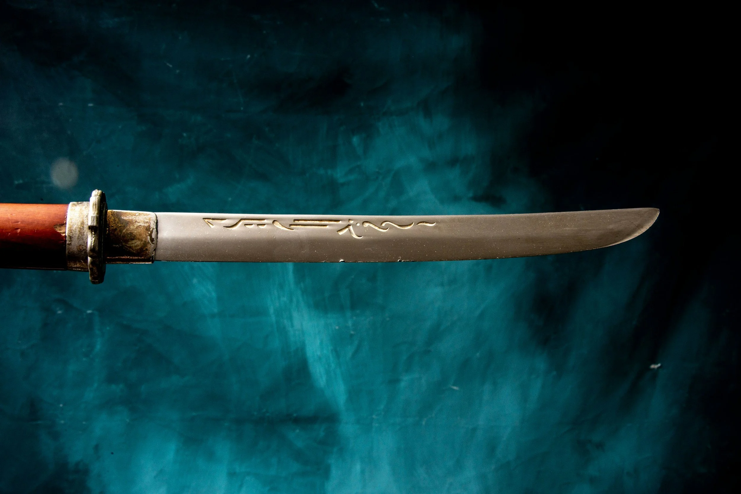 Close-up of a traditional Japanese katana sword with a wooden handle, metal guard, and engraved blade against a dark background.