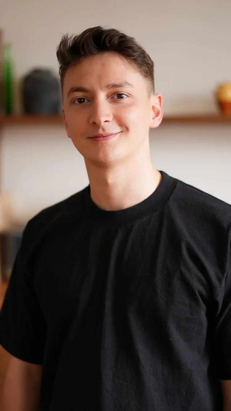 A young man with short brown hair smiling, wearing a black T-shirt, standing indoors with a blurred background that includes shelves and decorative items.