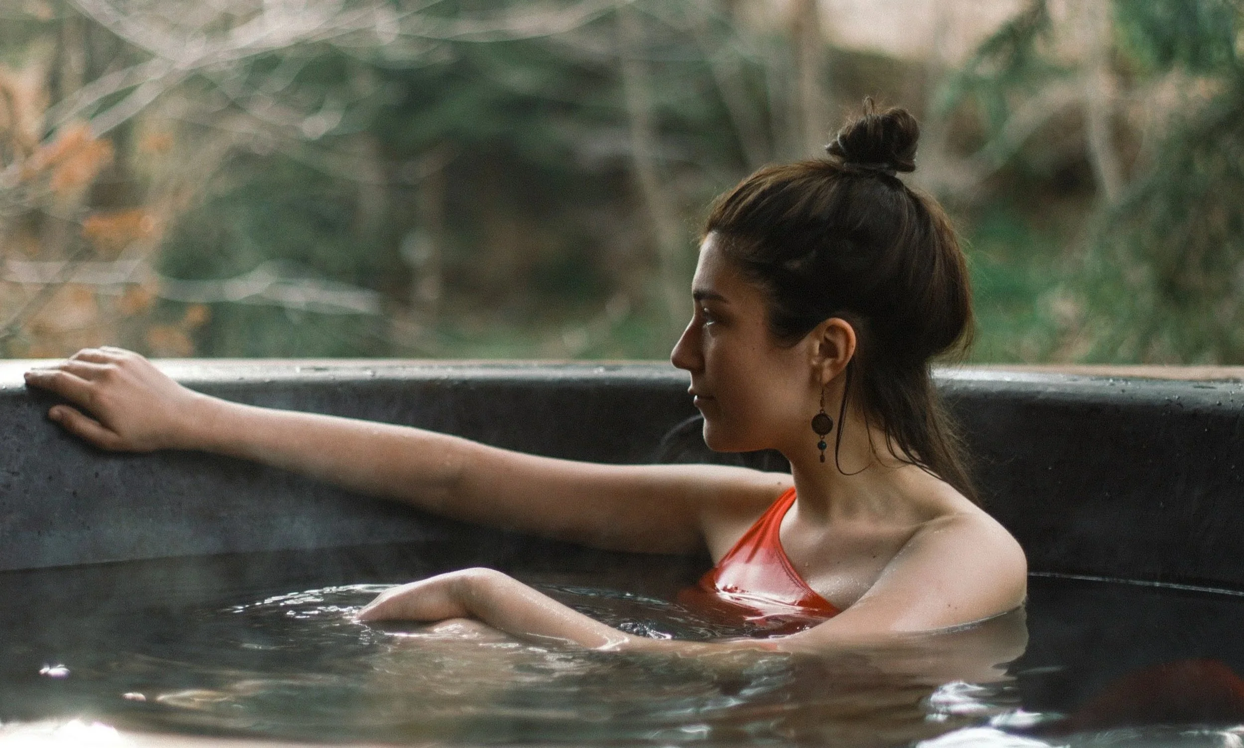 A woman with dark hair in a bun, wearing earrings and an orange top, is relaxing in a hot tub outdoors, with her arms resting on the edge and looking to the side, surrounded by a blurred natural background.