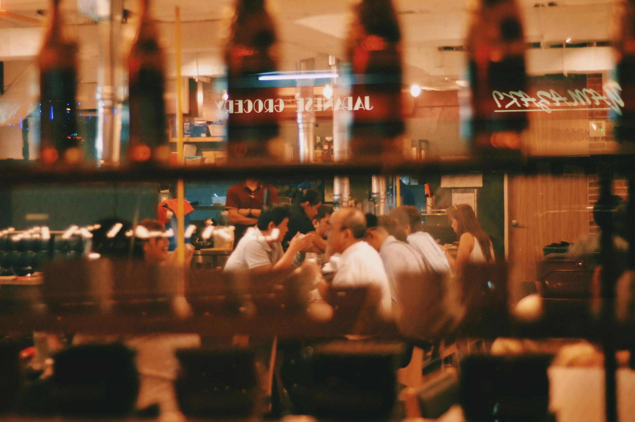 Scene inside a restaurant viewed through a blurry, glass window with vertical bars. People are sitting at tables, dining and talking. The background shows a sign that reads 'JAPANESE FOOD'.