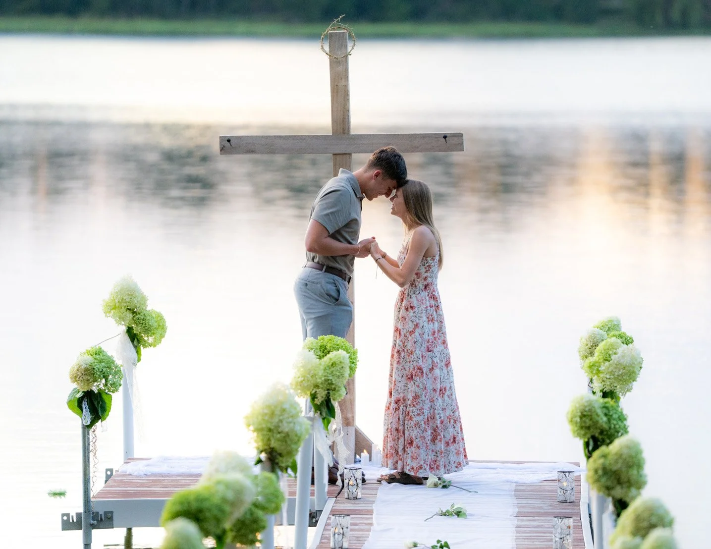 A couple getting married outdoors on a dock by the water, holding hands with foreheads touching, surrounded by floral decorations.
