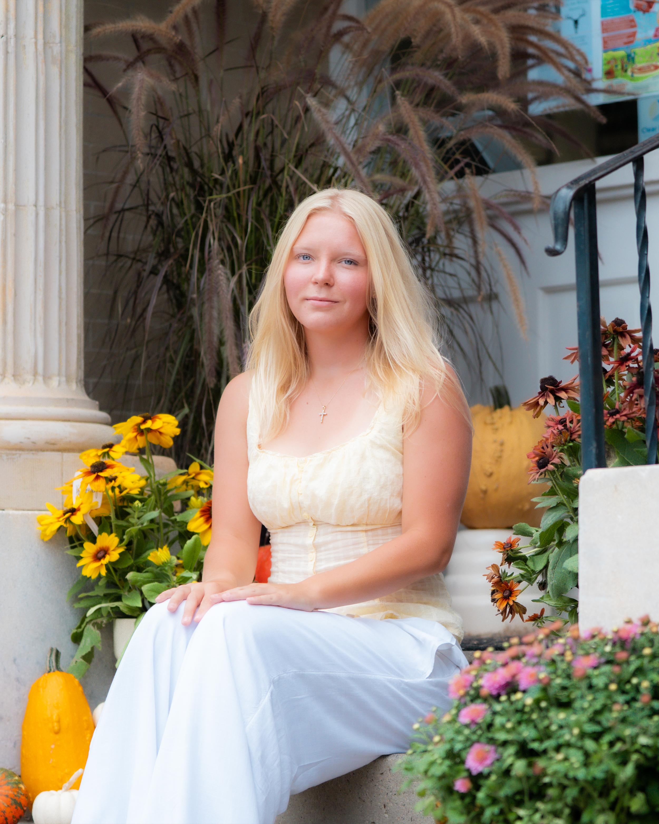 A young woman with long blonde hair and fair skin sitting outdoors surrounded by fall flowers and pumpkins, with a black metal railing and autumn plants. She is wearing a sleeveless yellow dress with a cross pendant necklace.