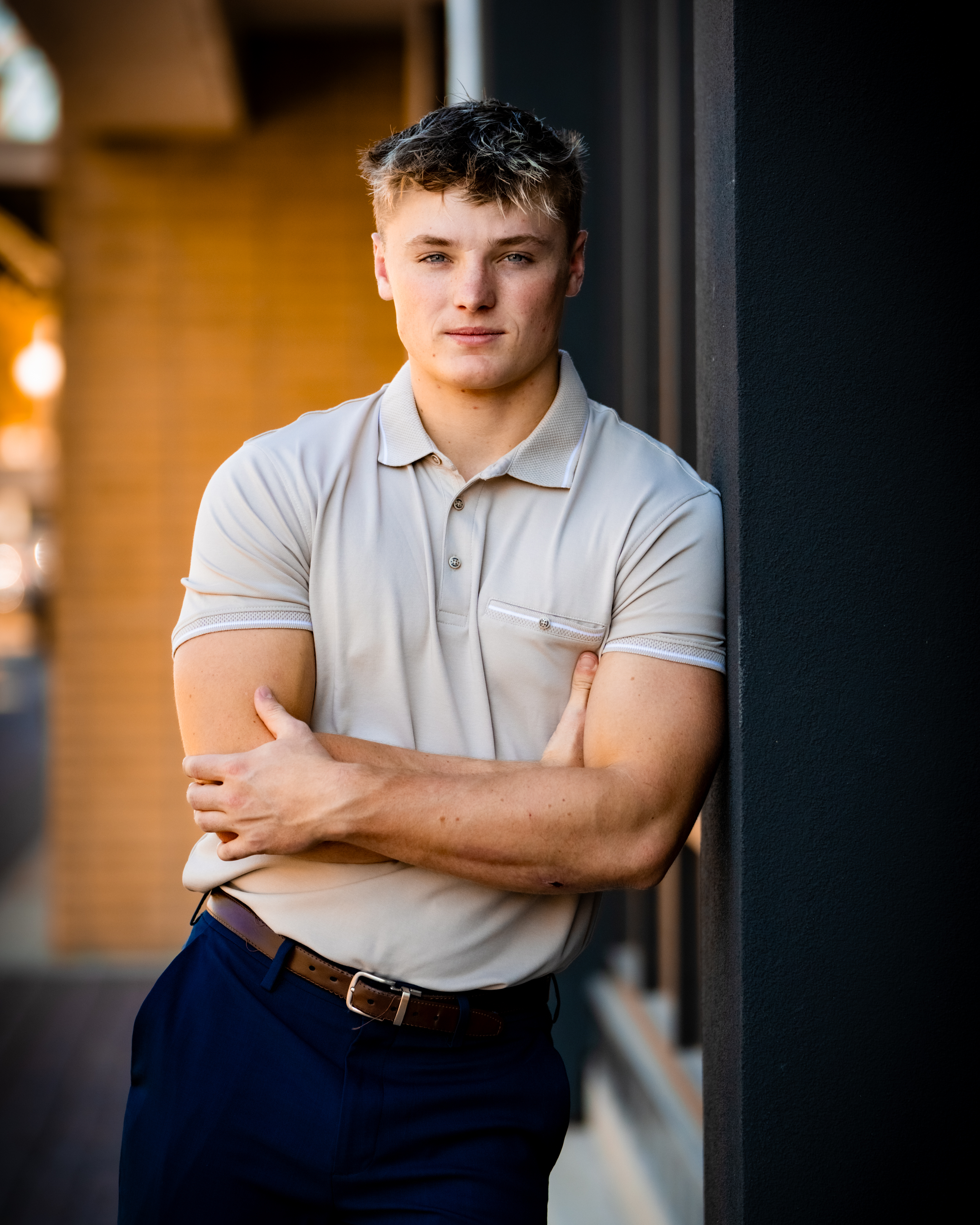 A young man with blond hair and blue eyes standing against a dark wall, wearing a light beige polo shirt and navy blue pants with a brown belt, arms crossed, looking at the camera.