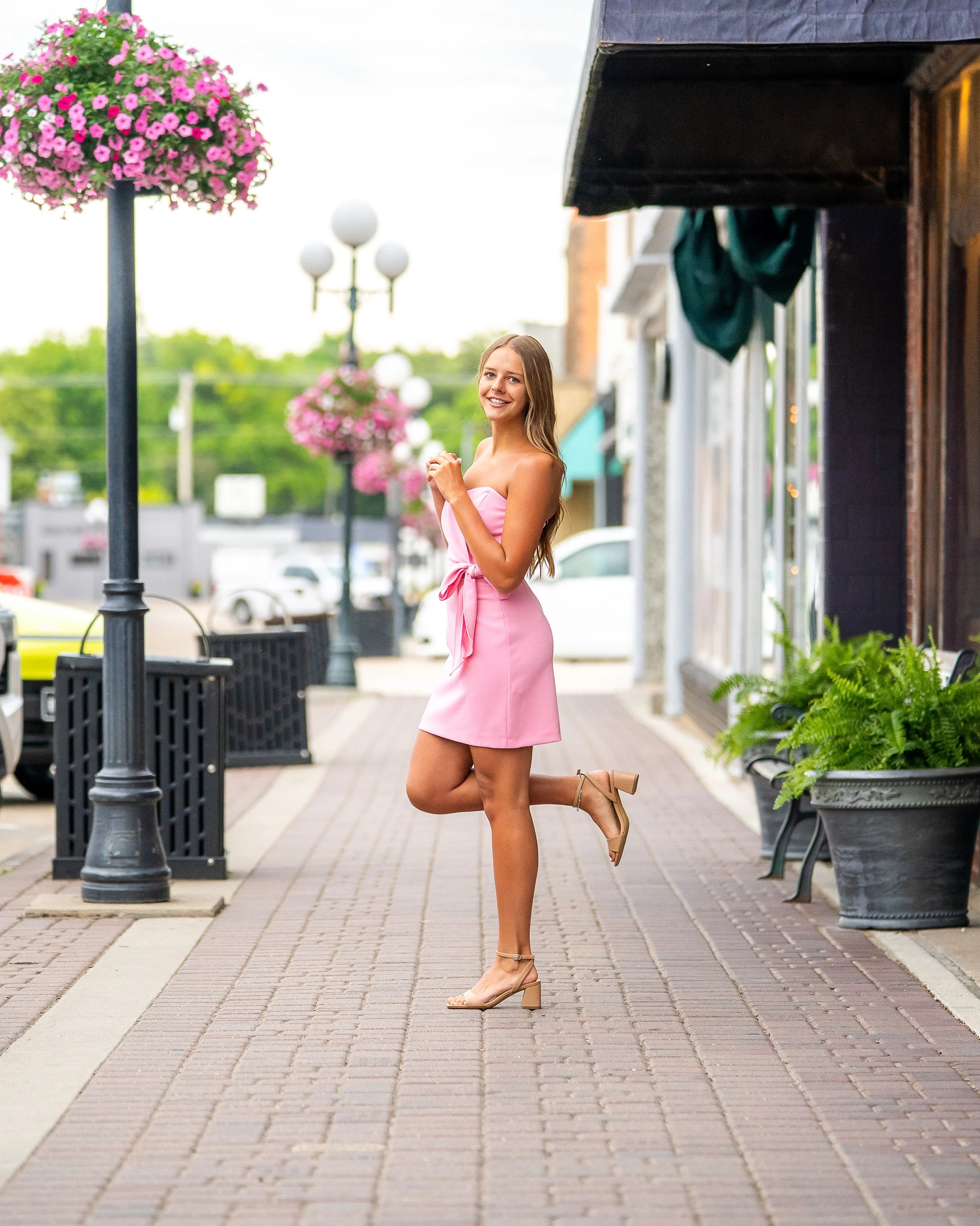 woman in pink dress and beige heels kneeling on a brick sidewalk with hanging flower baskets and street lamps in the background.