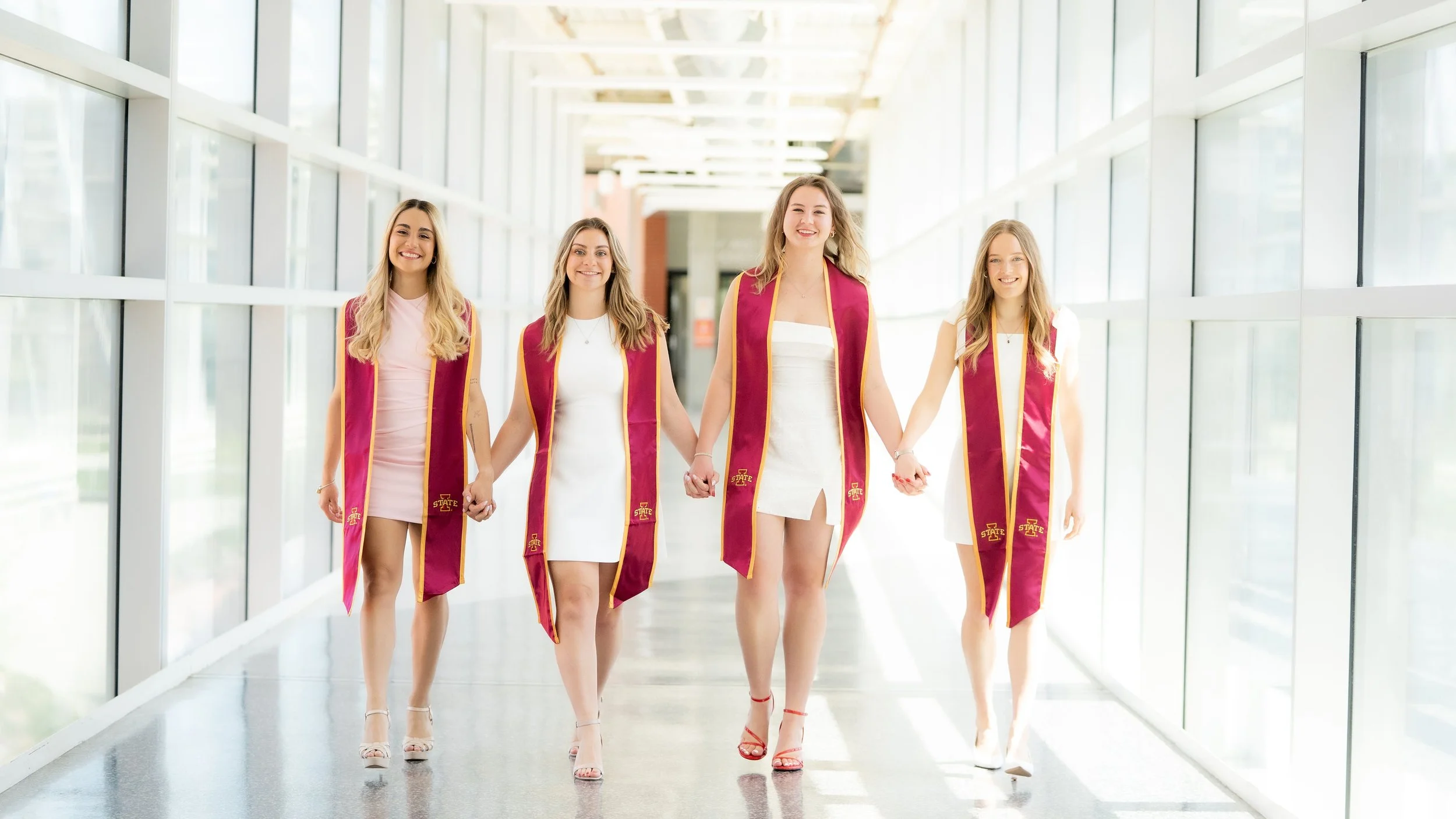 Four young women in white dresses wearing maroon and gold graduation stoles walking hand-in-hand through a bright, modern corridor with large windows.
